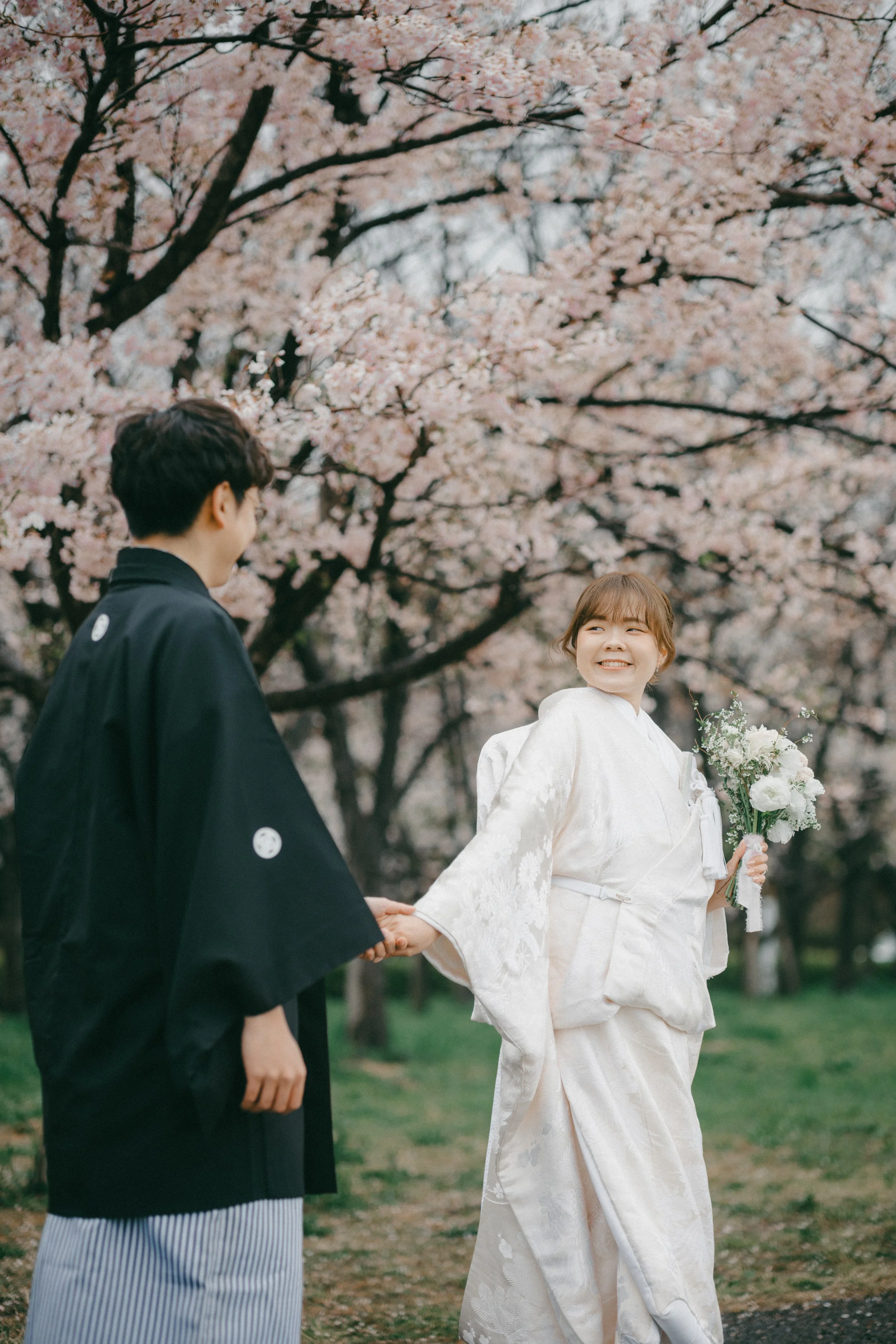 A bride in a white kimono holding a bouquet of white flowers and a groom in a black kimono smiling and holding hands under cherry blossom trees.