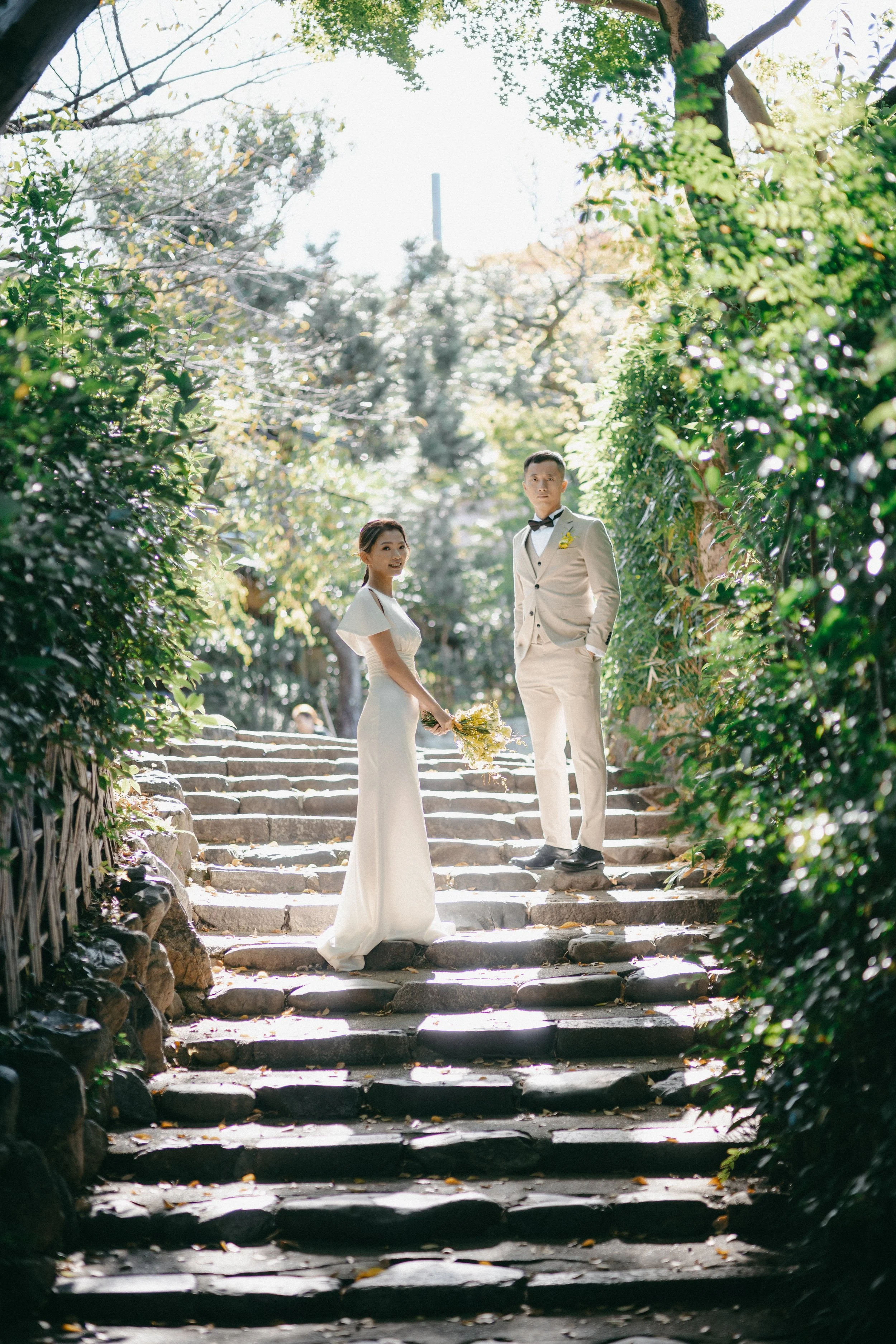 A bride and groom stand on stone steps in a lush, green outdoor setting during a wedding. The bride is holding a bouquet of yellow flowers and smiling, while the groom stands beside her in a light-colored suit with a black bow tie, looking into the c