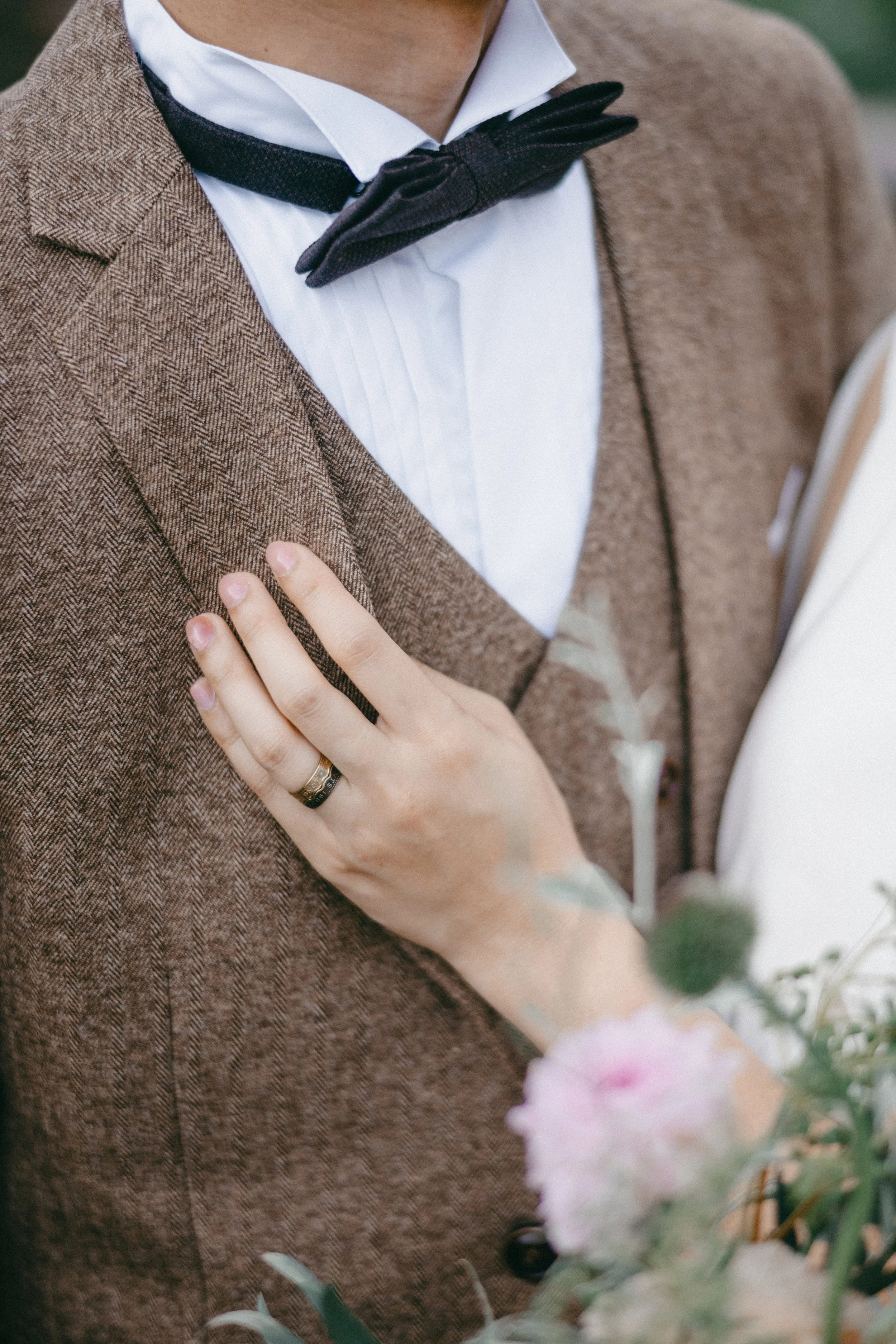 Close-up of a man in a brown suit and white shirt, wearing a black bow tie, with a woman's hand on his chest, showing a wedding ring, and some pink flowers in the foreground.