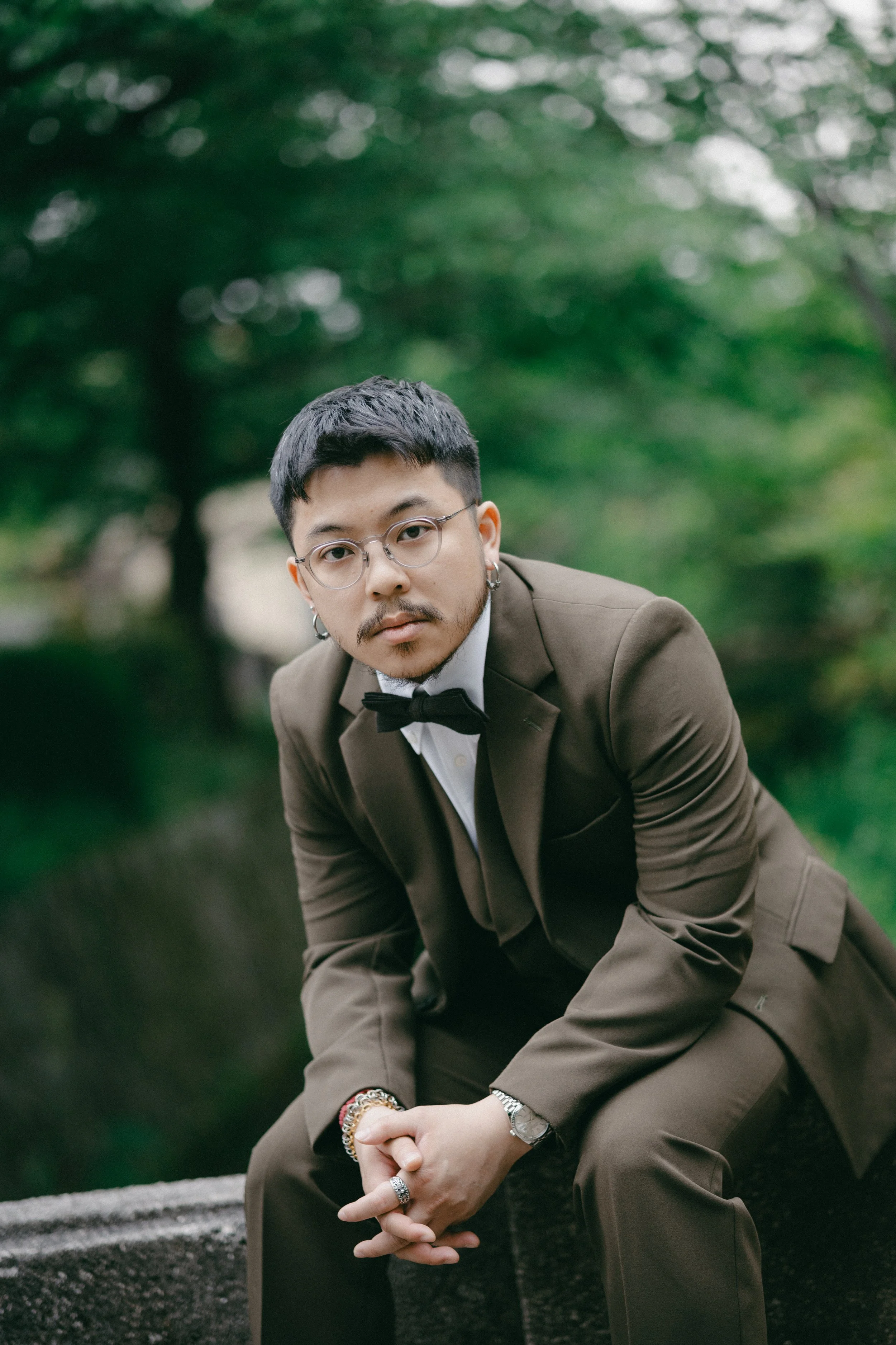 A man in a brown tuxedo with a bow tie and glasses sitting outdoors with green trees in the background.
