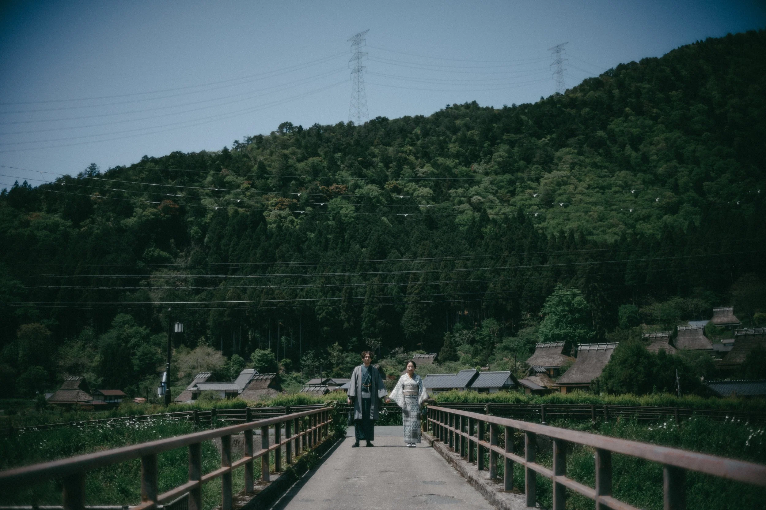 Two people in traditional Japanese clothing walking on a bridge in a rural village with mountain and forest in the background.
