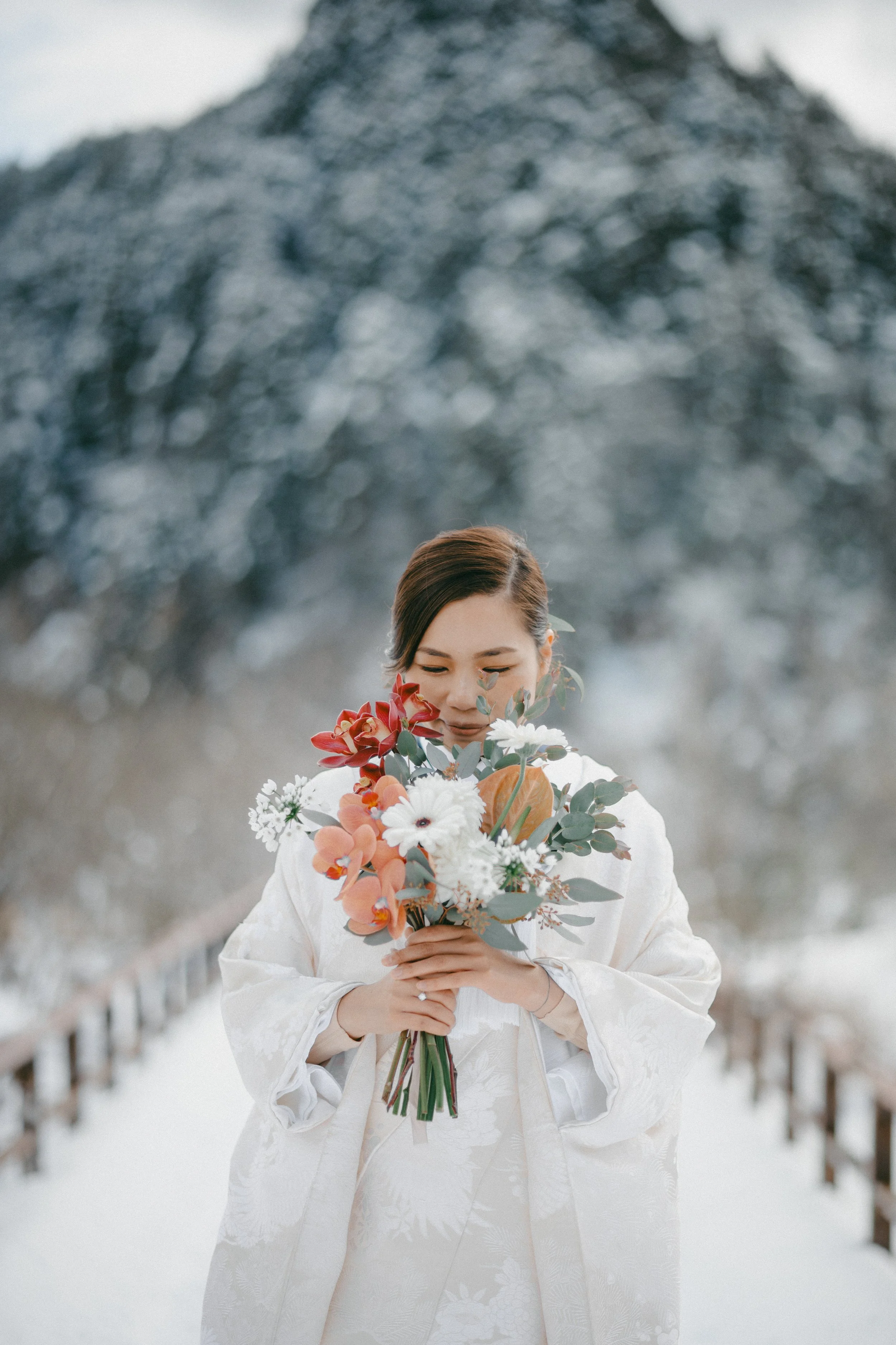 A woman in a wedding dress holding a bouquet of flowers outdoors in a snowy landscape with mountains in the background.
