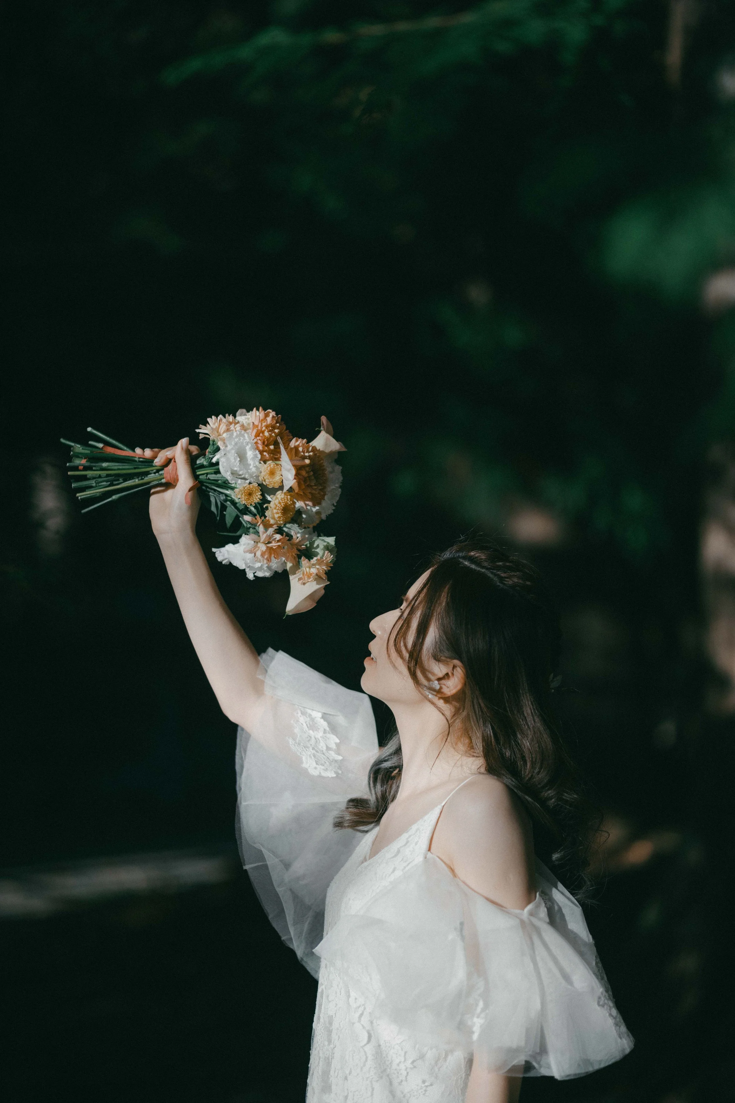 A woman in a white dress holding a bouquet of flowers in an outdoor setting with dark, blurry background.