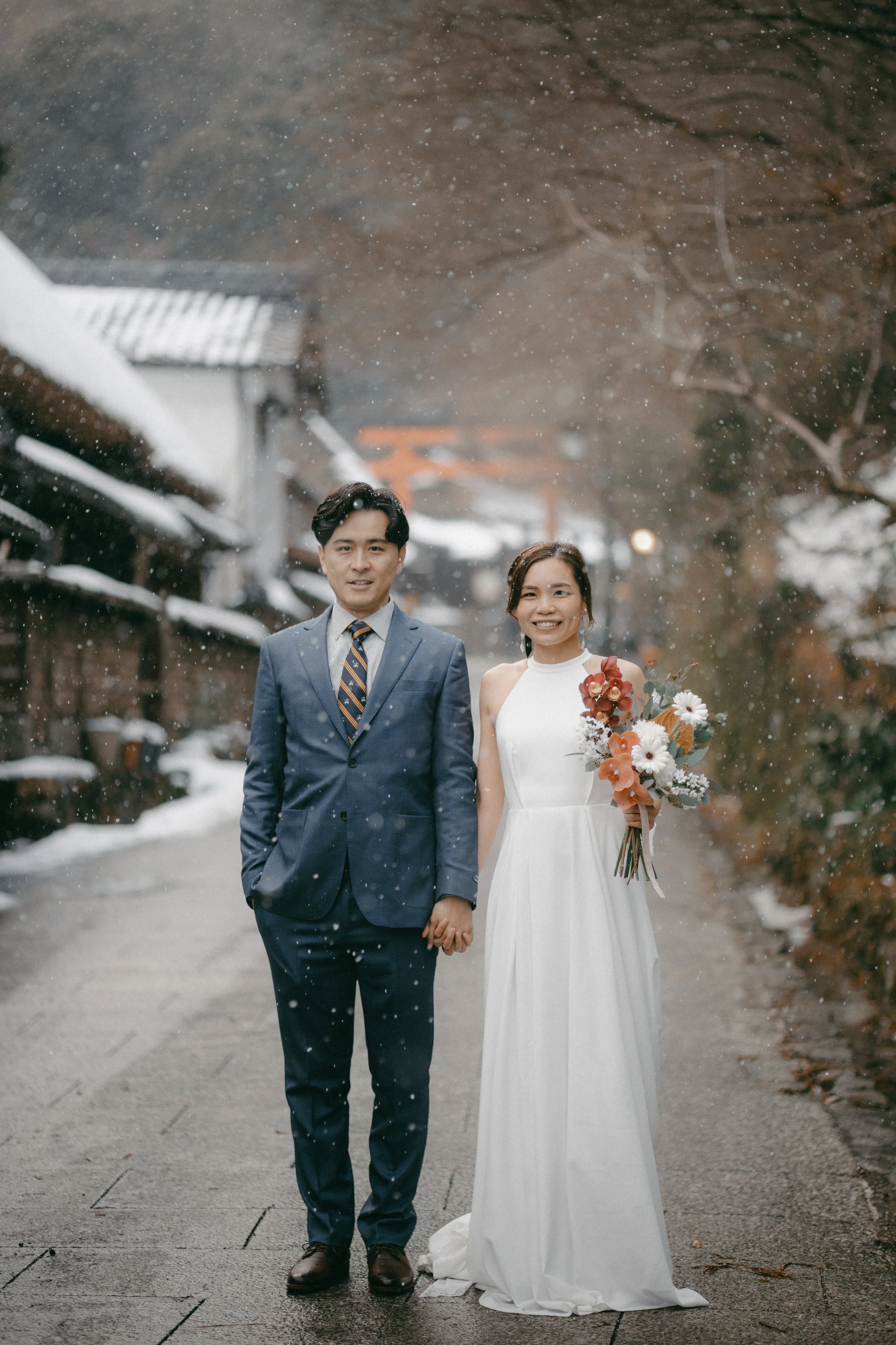 A newlywed couple stands hand-in-hand outdoors in a snowy setting, with traditional Japanese torii gate in the background. The bride is in a white wedding dress holding a floral bouquet, and the groom is in a navy suit with a striped tie.
