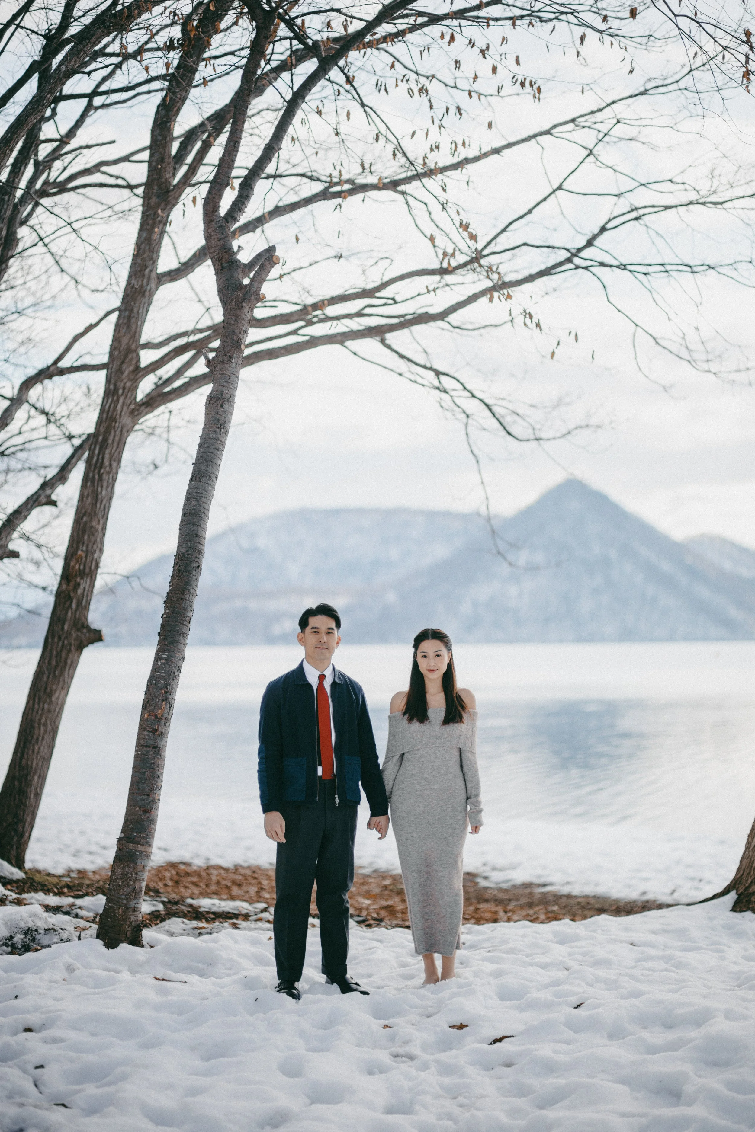 A man and woman standing hand in hand on snow-covered ground near a lake with snow-capped mountains in the background, trees with bare branches overhead.
