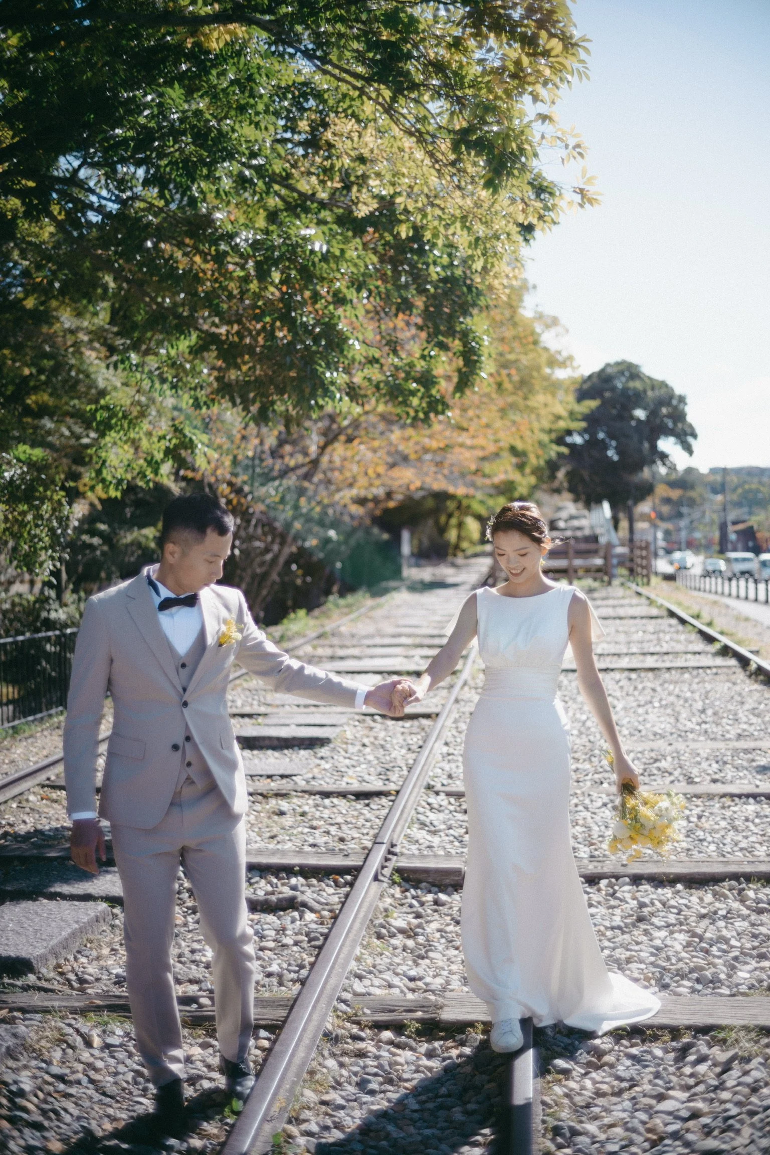 A newlywed couple walking hand-in-hand along train tracks outdoors on a sunny day, with trees and cars in the background.
