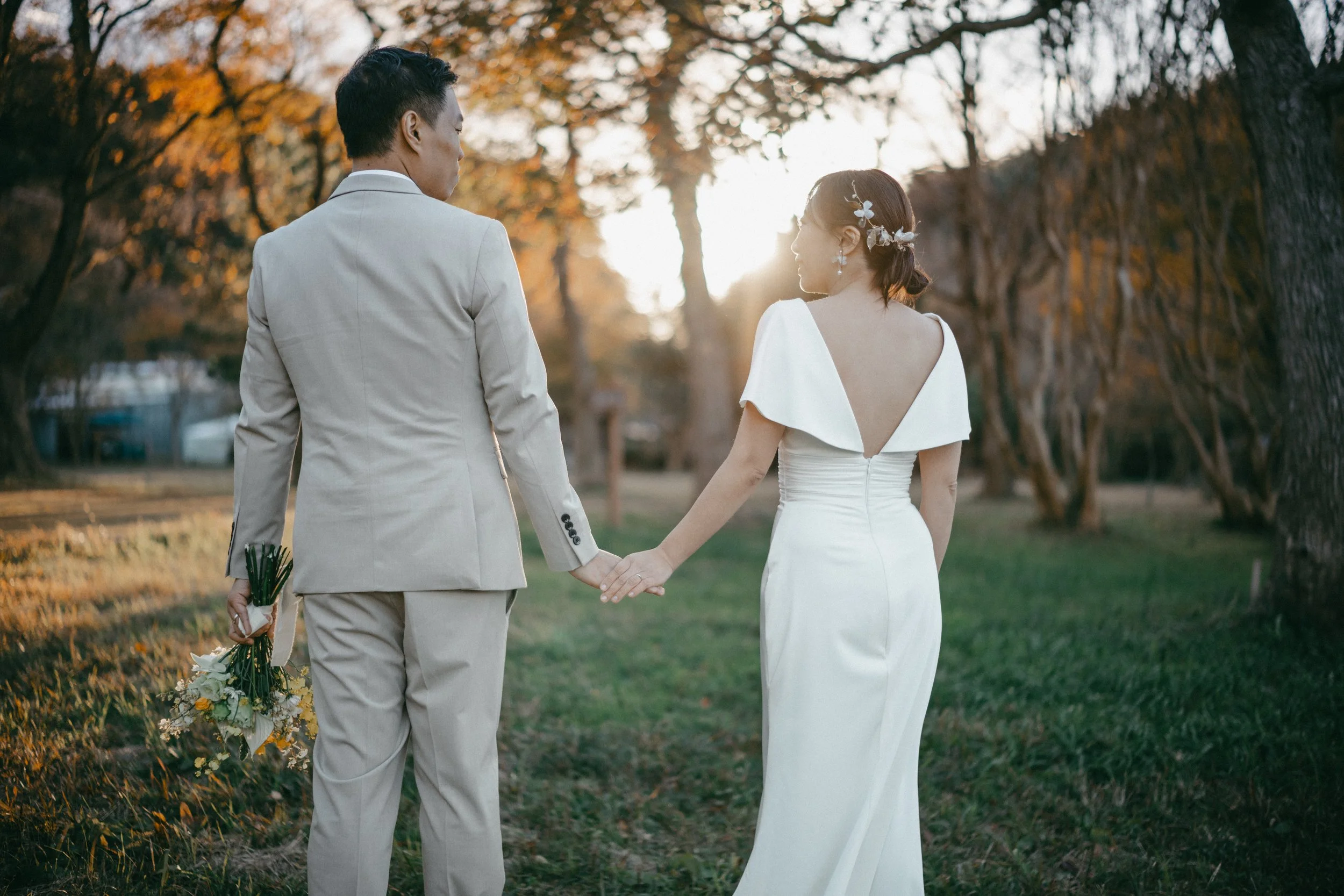 A bride and groom holding hands and walking in a park during sunset, dressed in wedding attire with trees in the background.