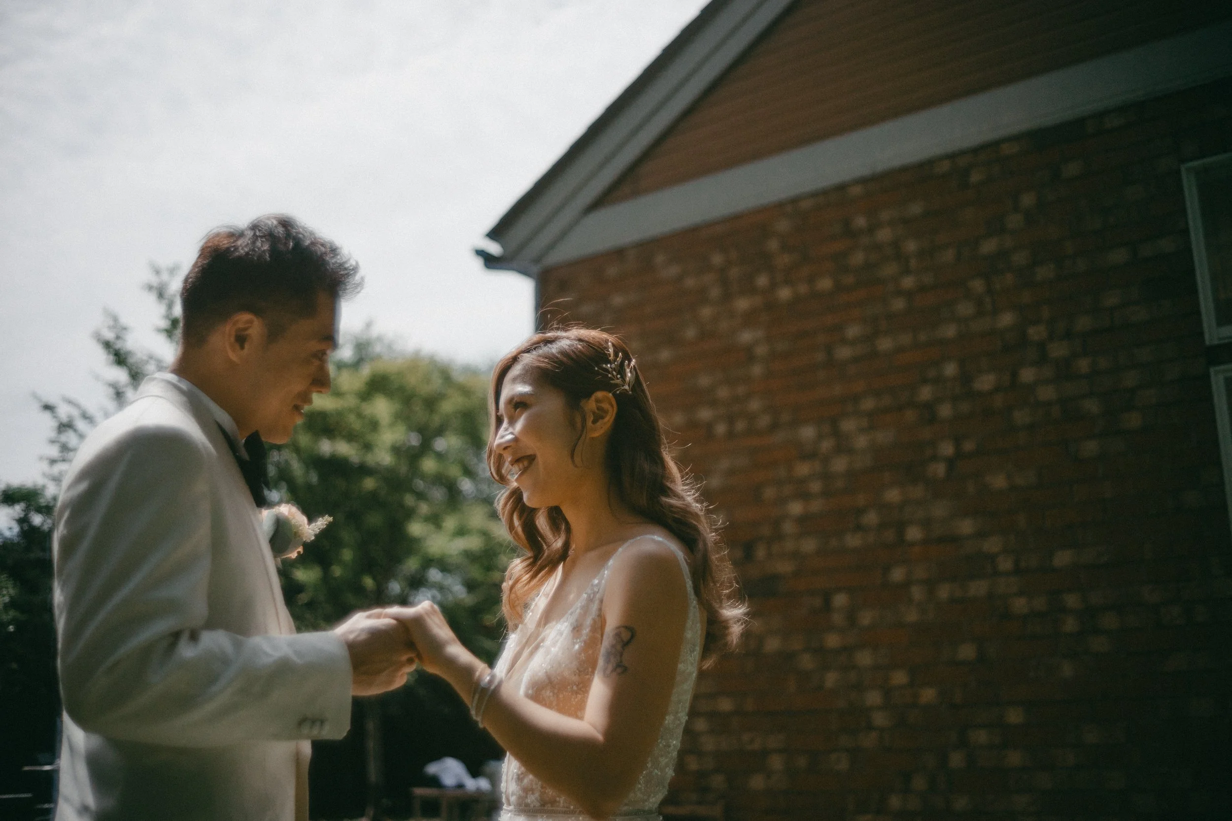 A bride and groom smiling and holding hands outside on a sunny day near a brick house.