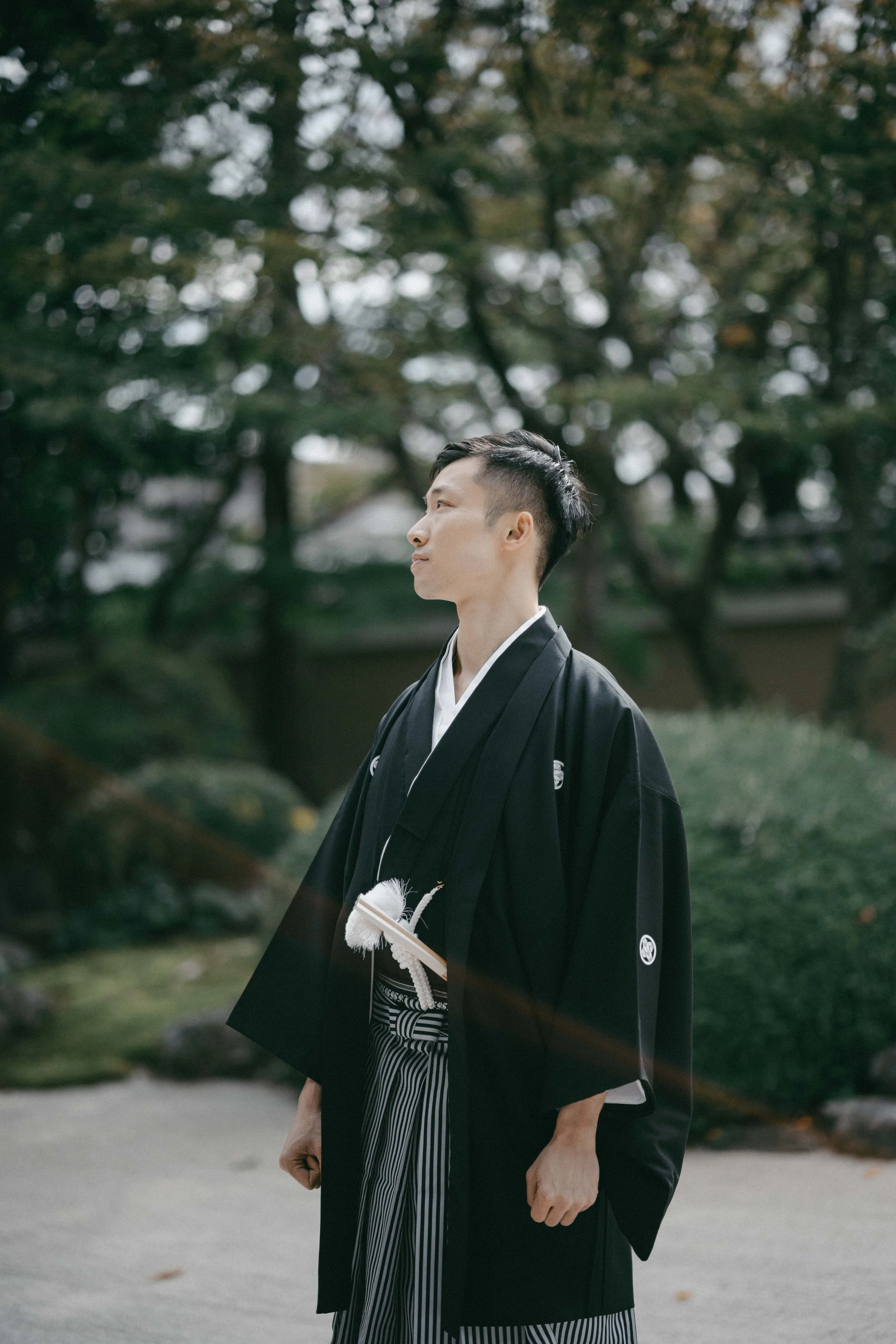 A young man in traditional Japanese attire stands outdoors in a garden with trees and bushes. He is wearing a black kimono with a white collar and striped hakama pants, holding a ceremonial fan.