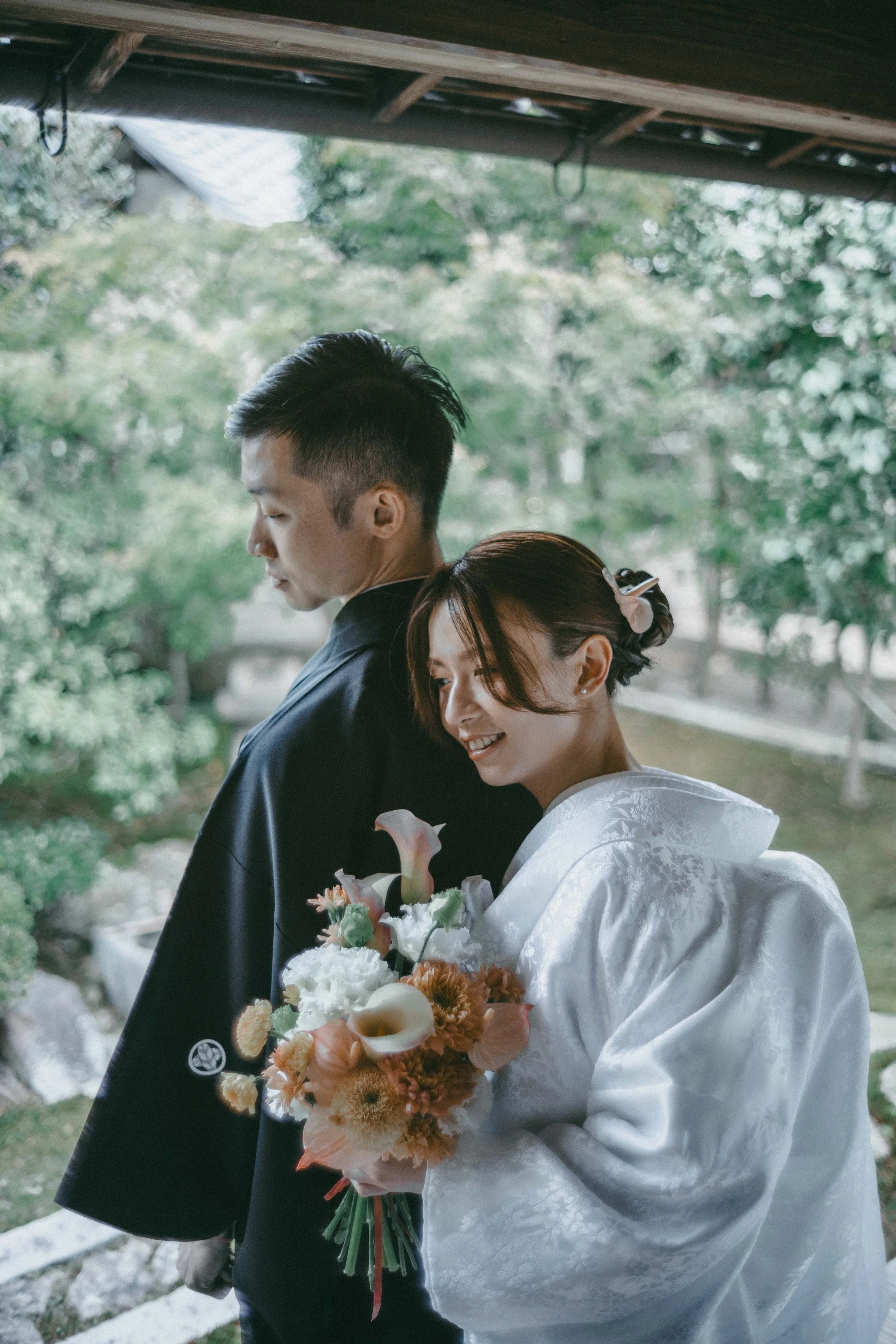 A bride and groom in traditional wedding attire outdoors, with the bride smiling and holding a bouquet of flowers, and the groom standing behind her with his head slightly tilted down.