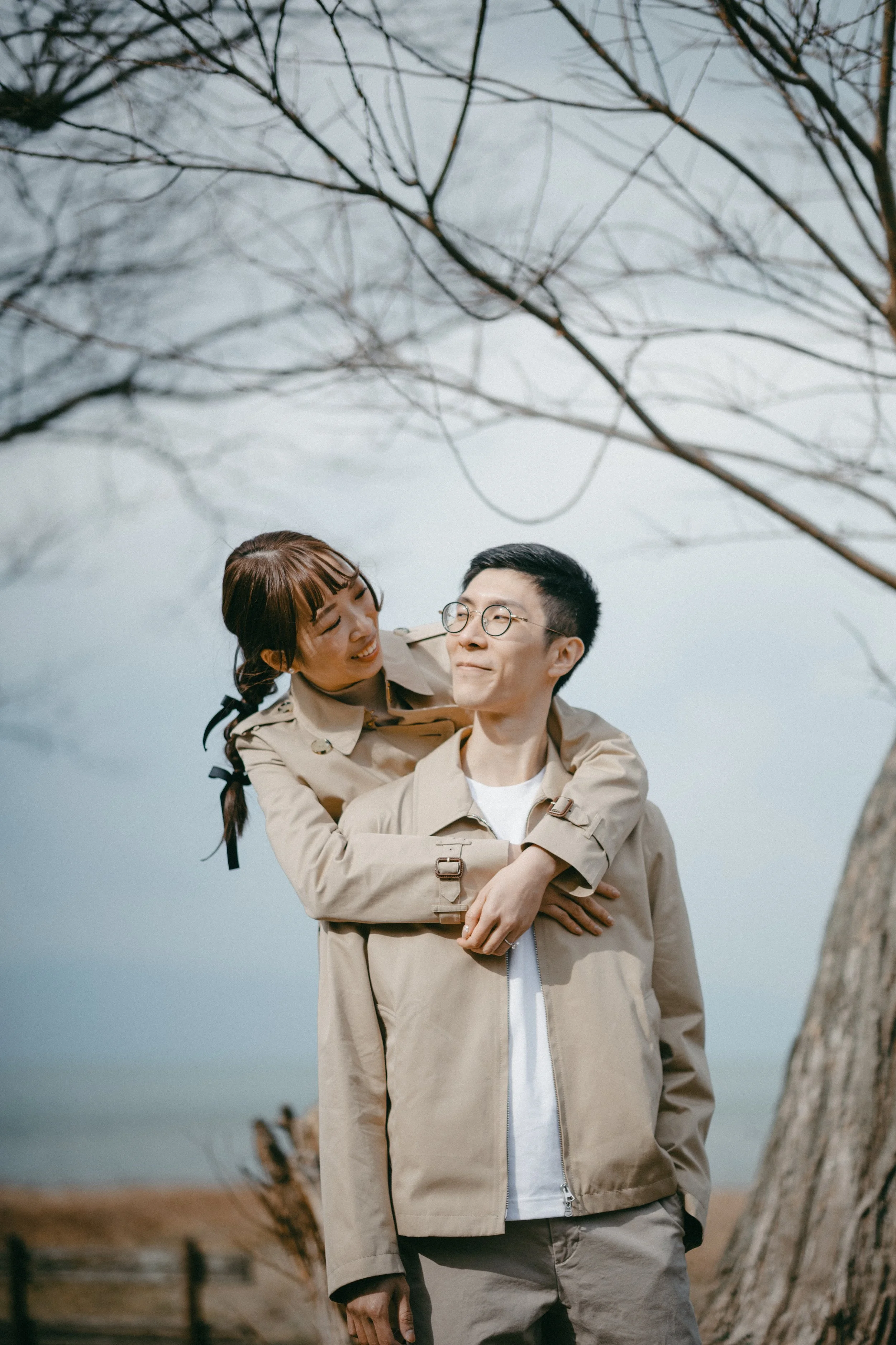 A couple wearing beige jackets standing outdoors on a cloudy day, with the woman playfully hugging the man from behind, both smiling, in front of leafless tree branches.