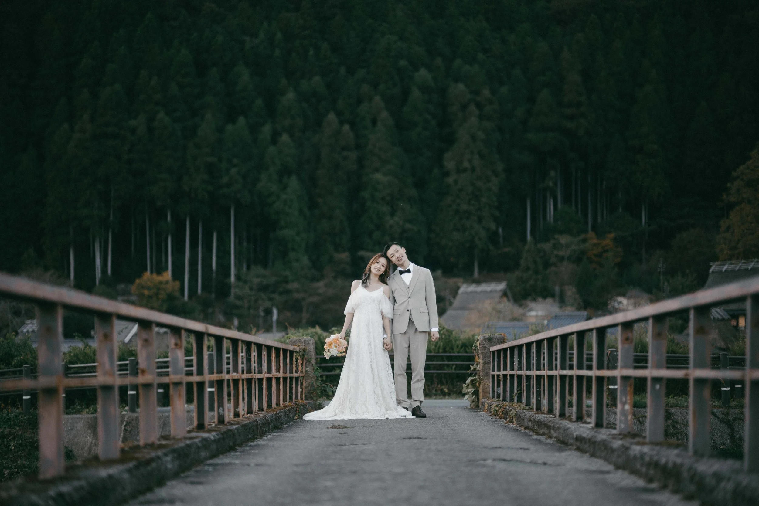 A bride and groom stand on a small bridge, holding hands and smiling, with a forested hillside in the background.