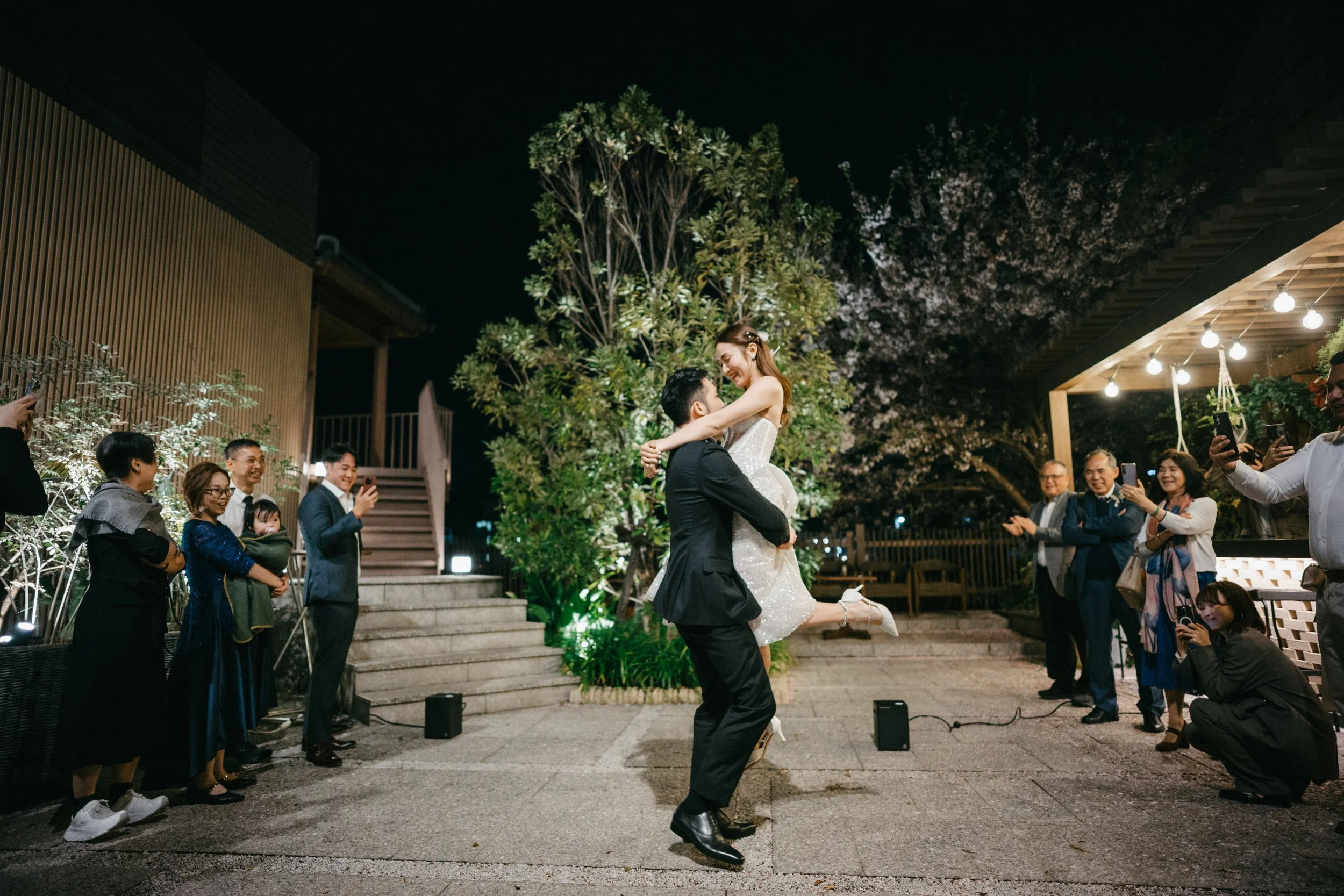 A couple dancing at a wedding reception outdoors at night, surrounded by friends and family who are taking photos and videos.