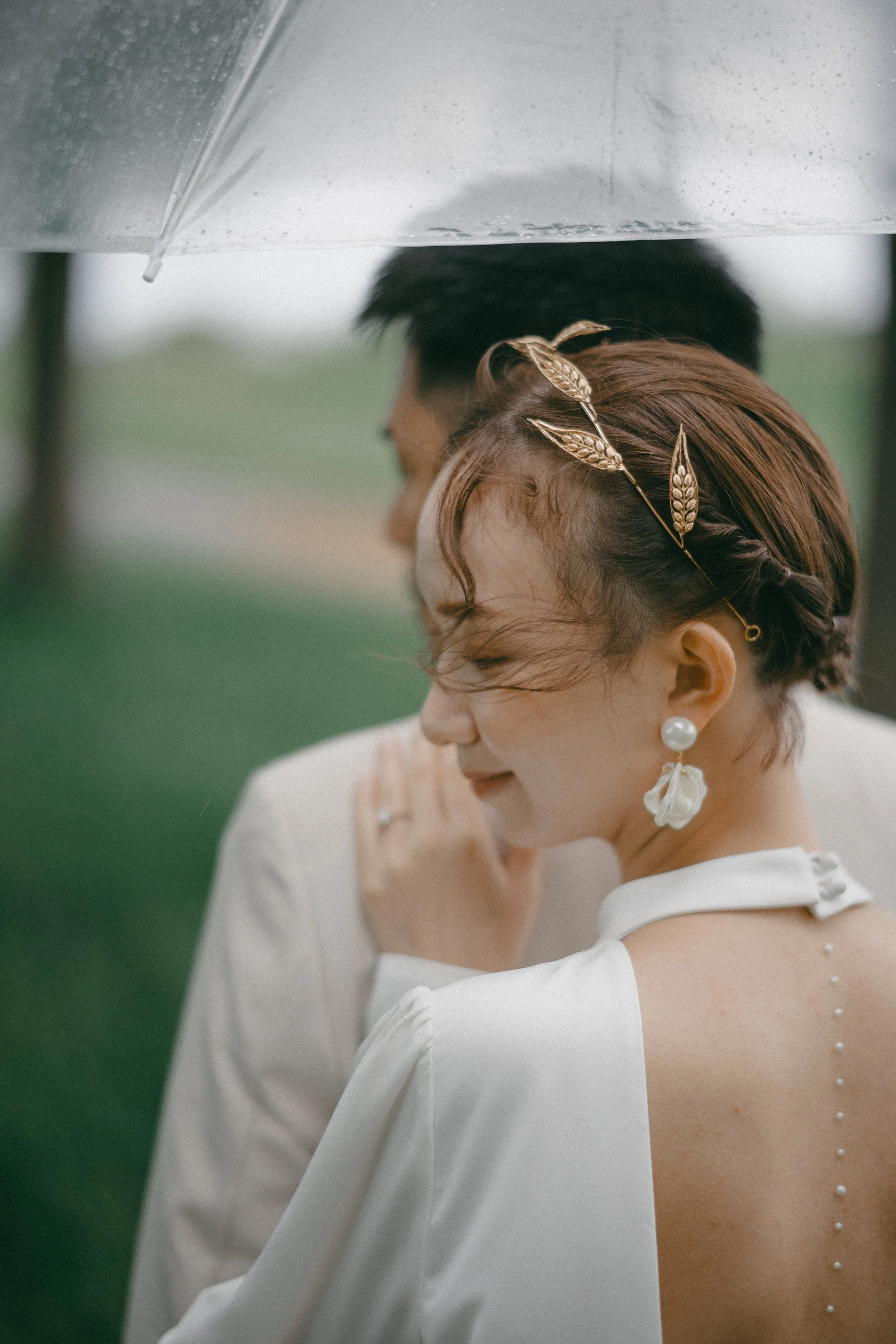 A woman and a man under a clear umbrella outdoors on a rainy day, with the woman wearing a white dress and gold leaf headband, and the man dressed in white.