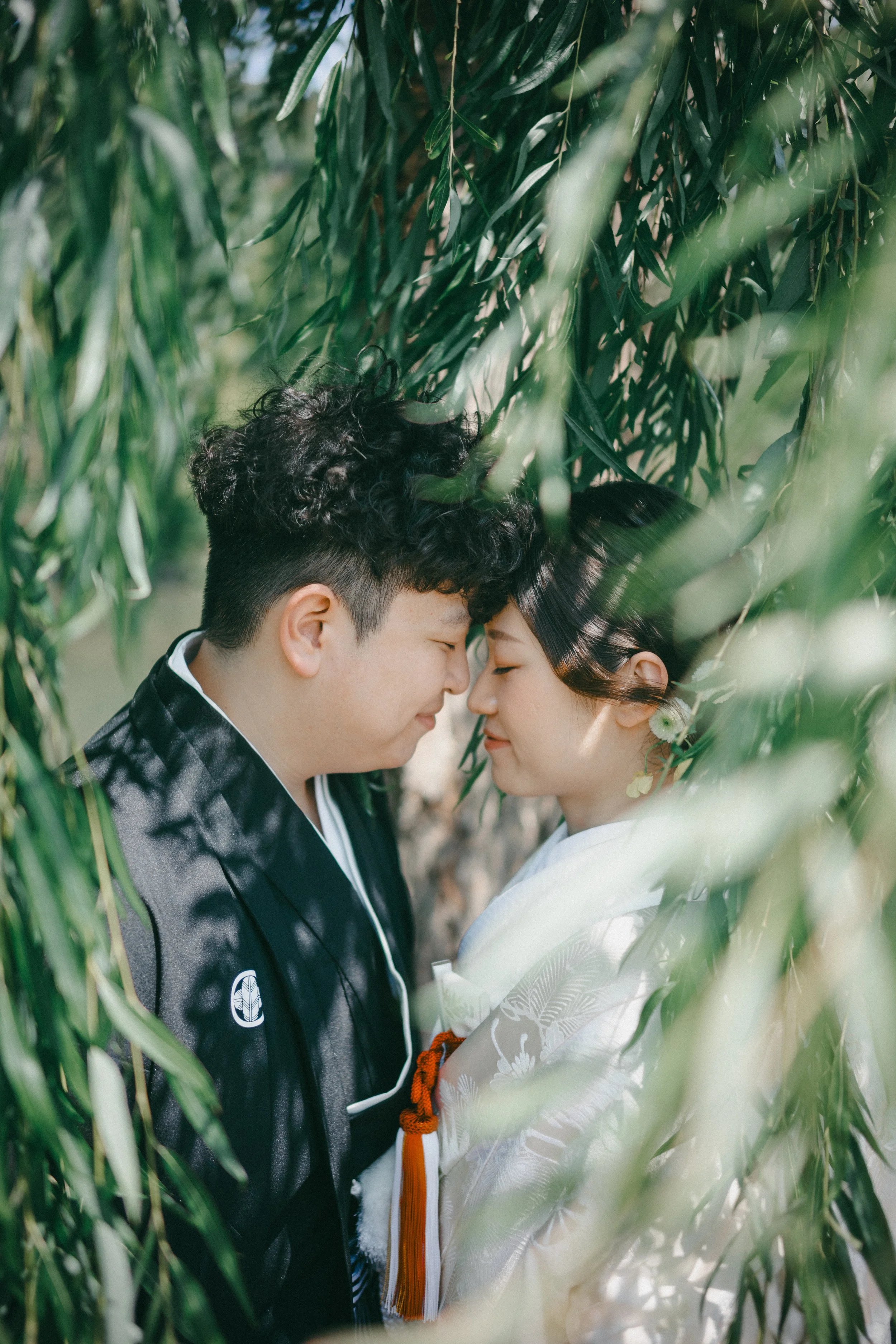 A couple dressed in traditional Japanese wedding attire sharing a tender moment with foreheads touching amidst green leafy trees.