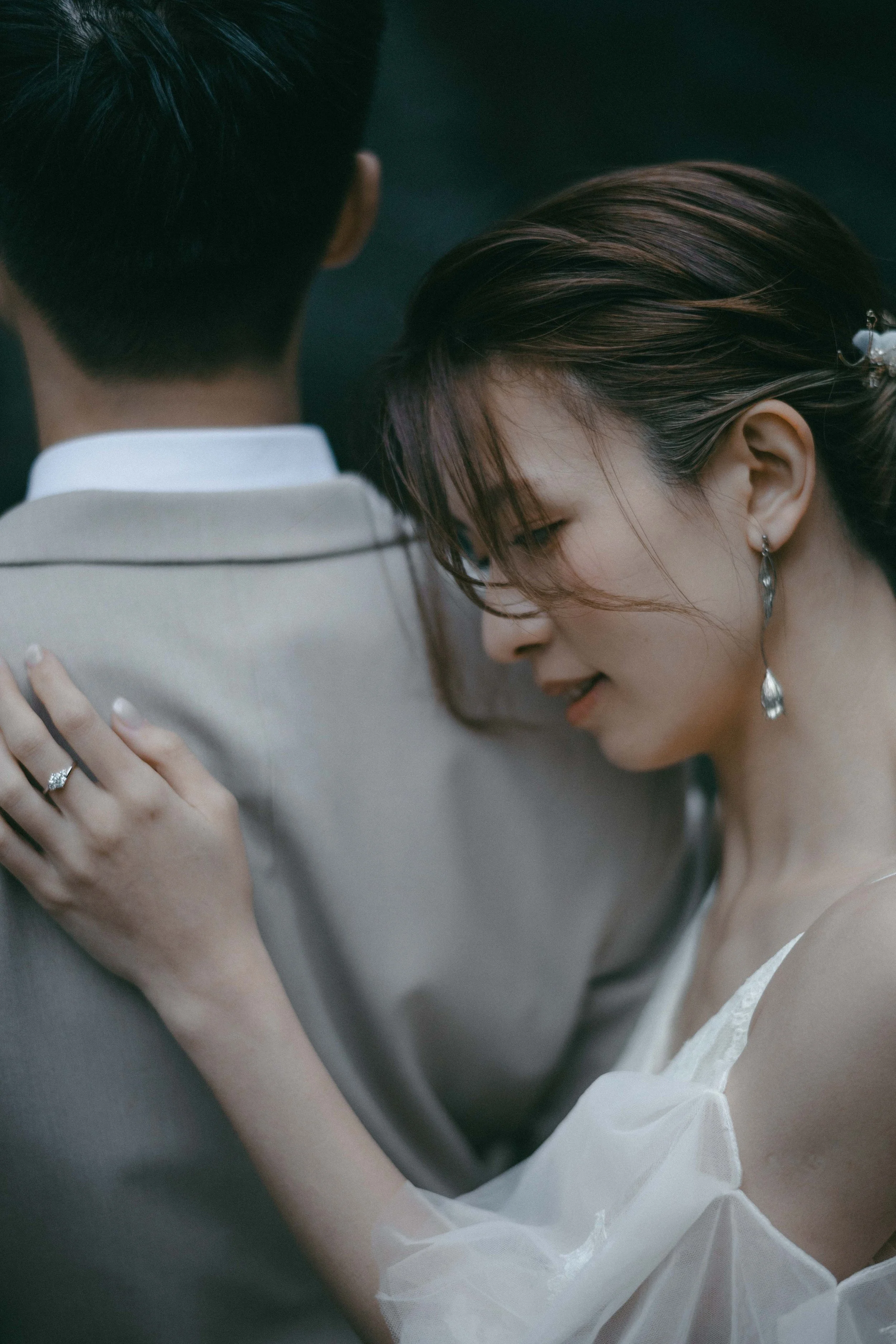 A woman with earrings and a white dress gently rests her hand on a man's shoulder, who is wearing a beige suit and white shirt, and has his face turned away.
