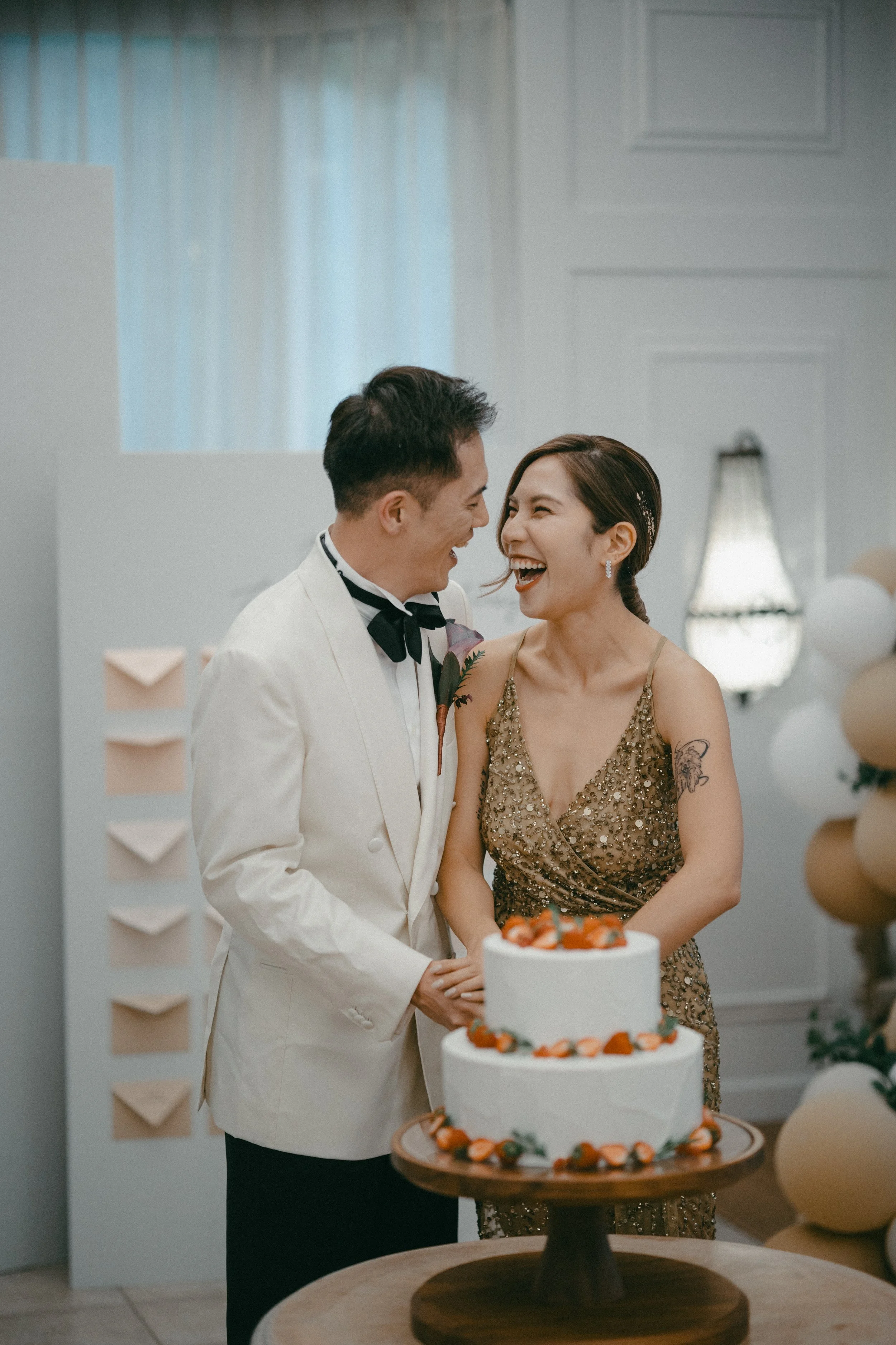 A couple dressed in formal attire is smiling and laughing while standing near a wedding cake at their celebration.