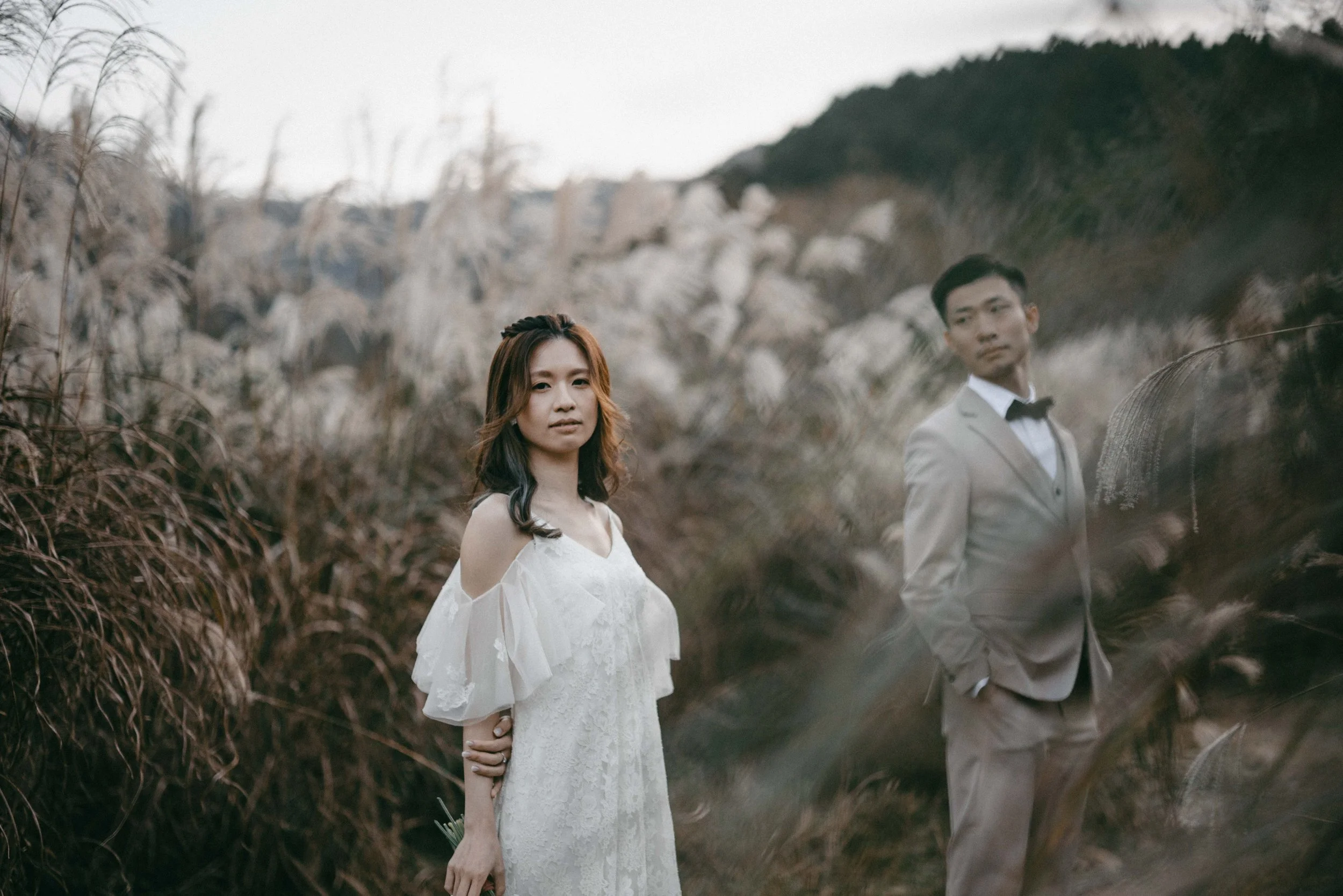 A woman in a white dress and a man in a gray suit with a black bow tie standing outdoors in tall grass with a mountain in the background.