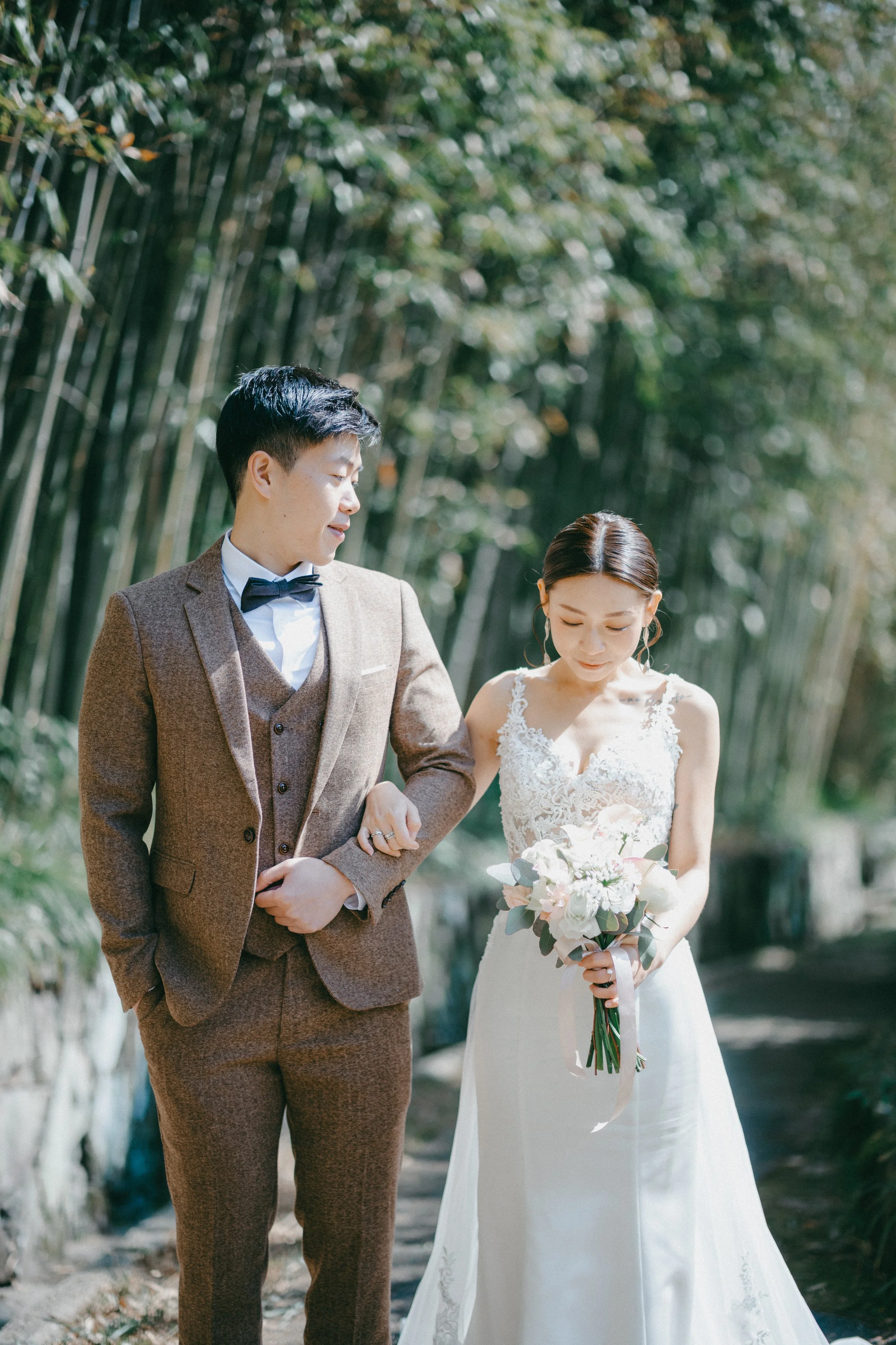 A bride and groom walk together outdoors, with the bride holding a bouquet of white flowers, in front of tall green bamboo stalks.