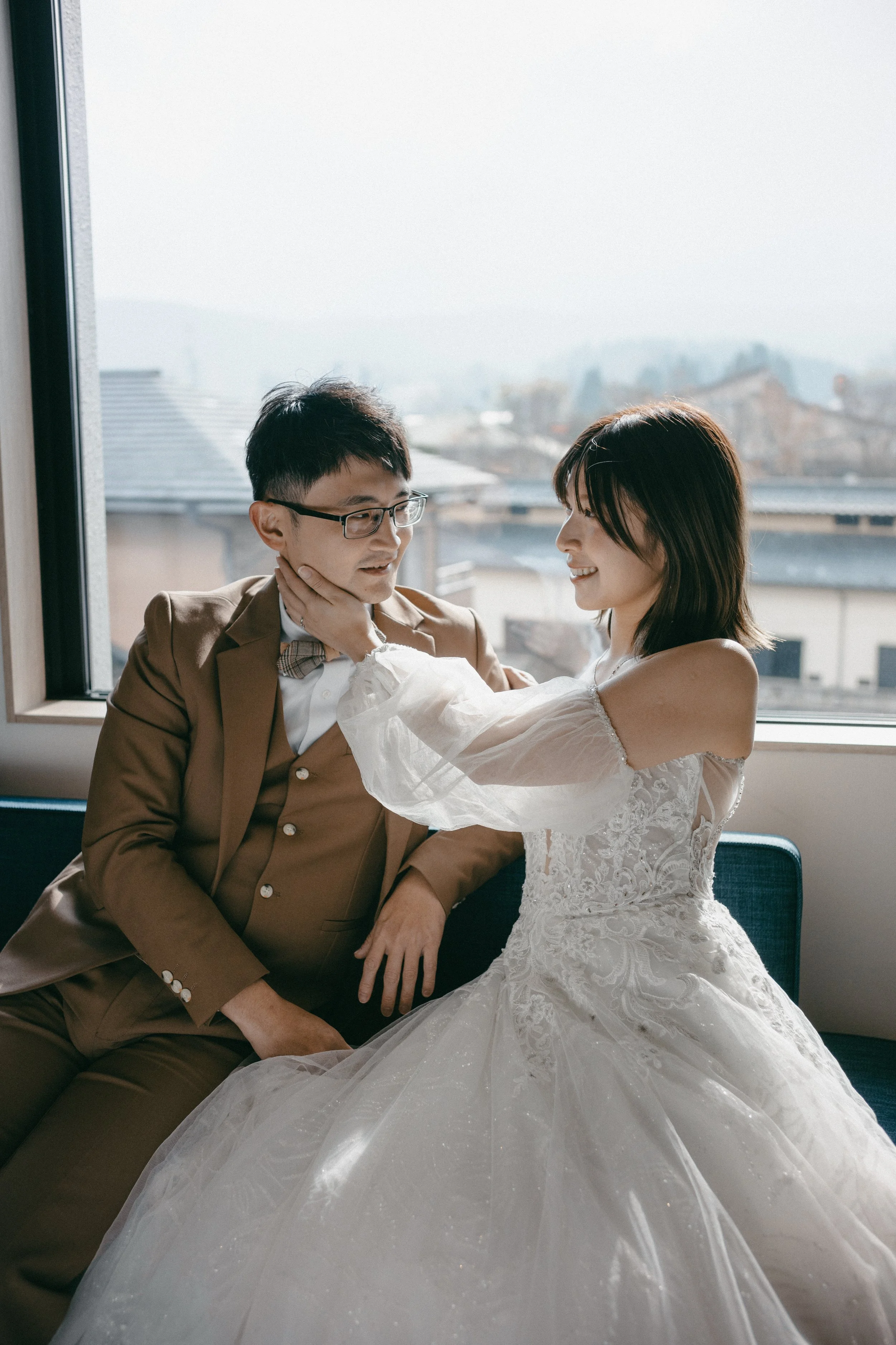 A bride and groom sharing a tender moment indoors near a large window, with the bride gently touching the groom's face, both smiling.
