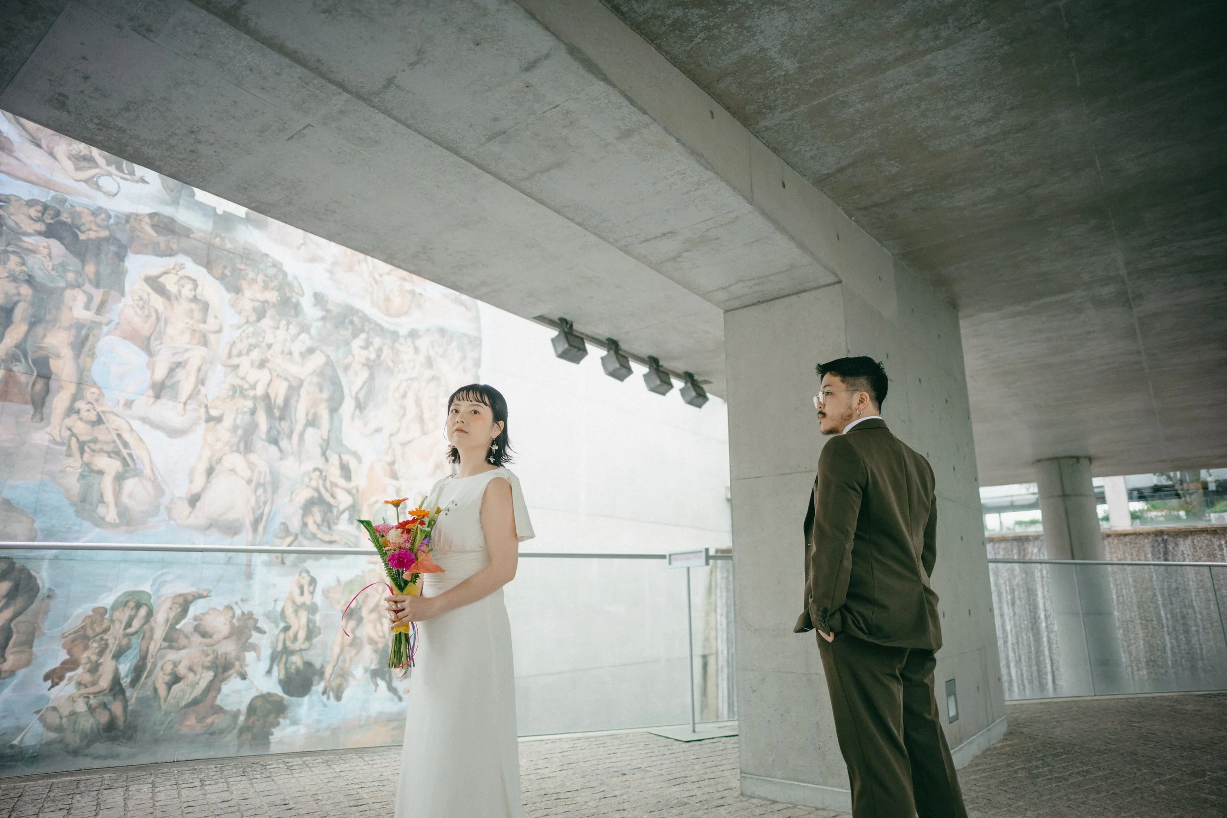 A woman in a white dress holding a bouquet of colorful flowers at an art gallery, with a man in a brown suit standing nearby, all in a modern, concrete interior.
