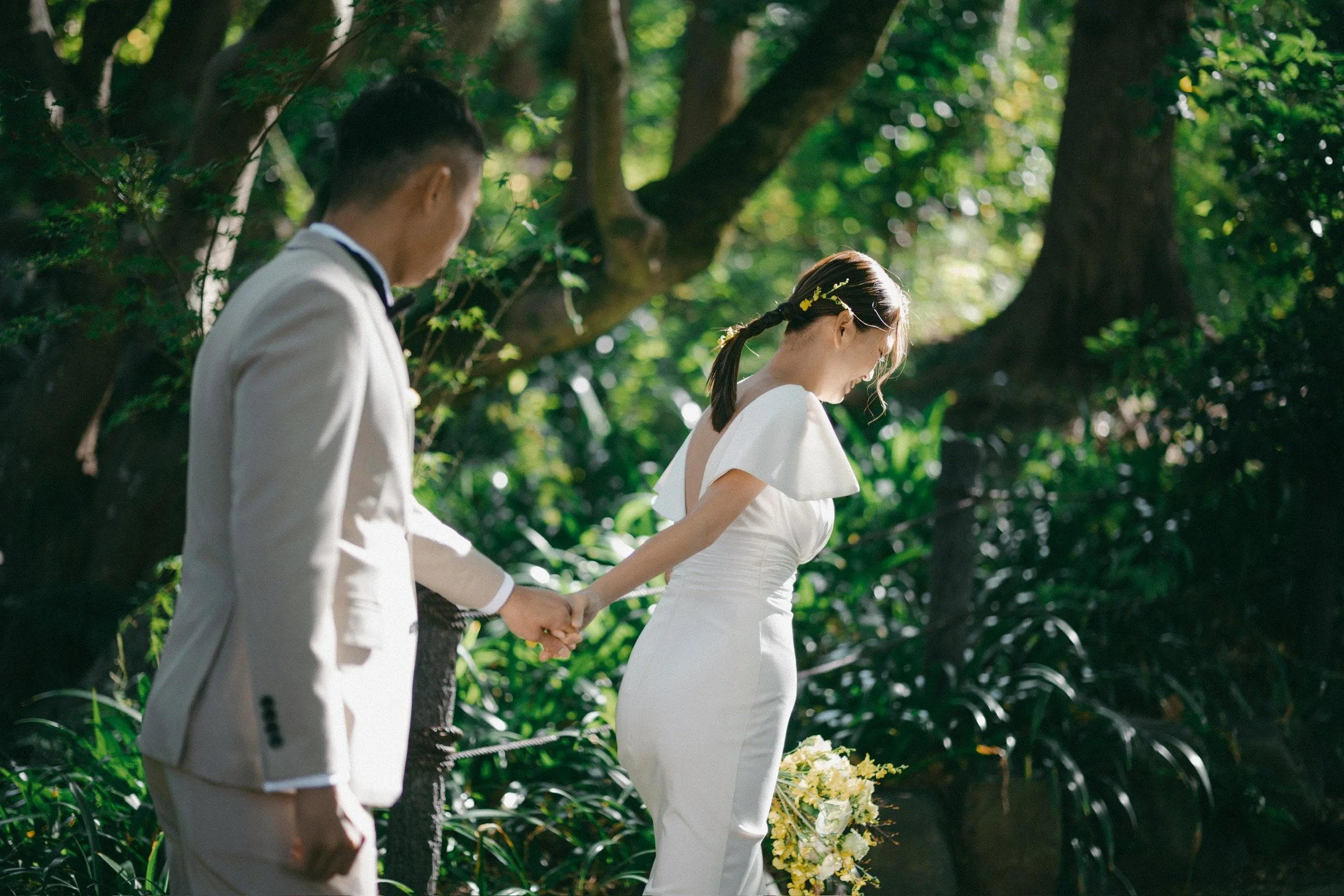 A bride and groom holding hands in a lush green outdoor setting, with sunlight filtering through the trees.