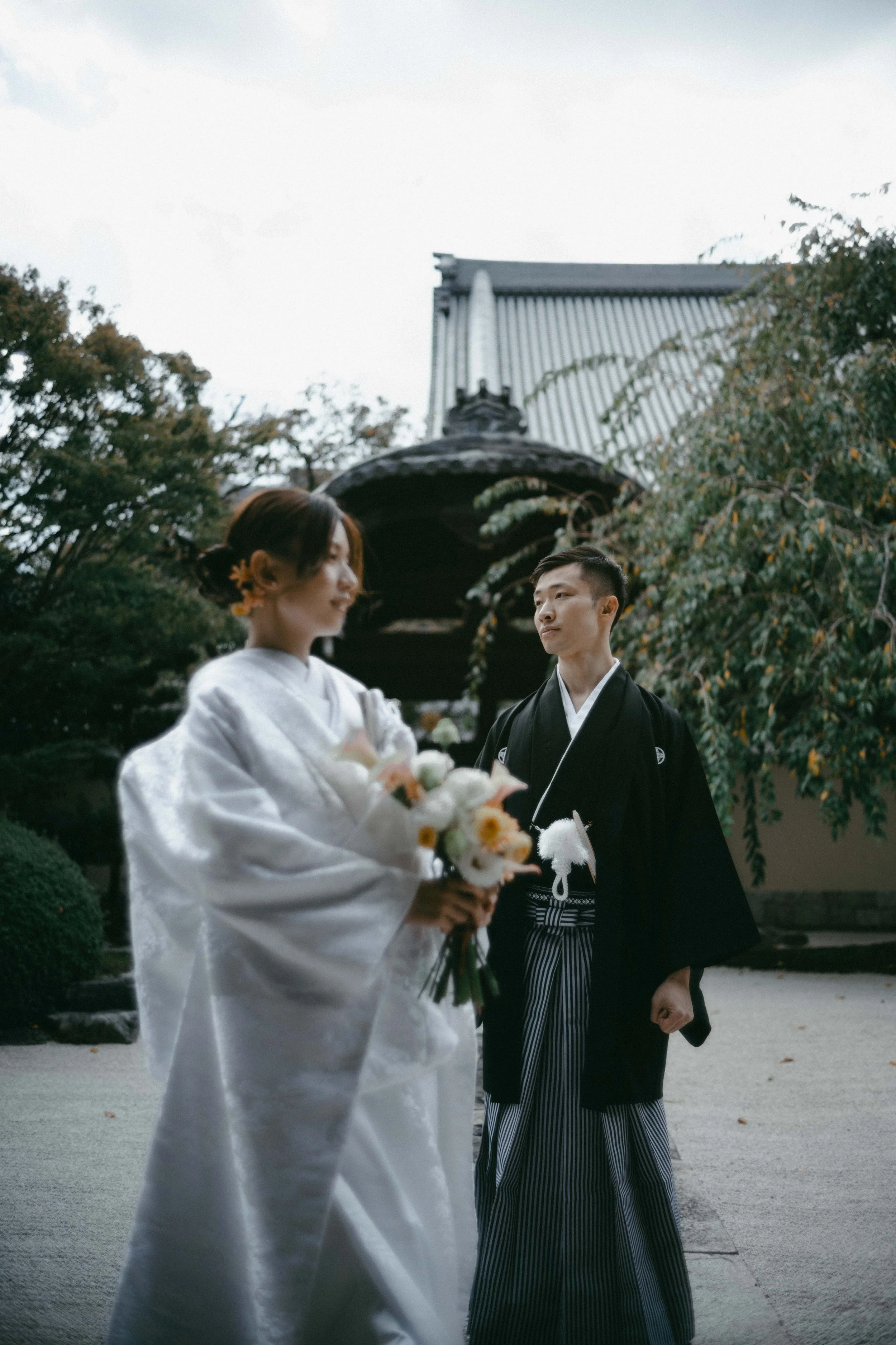 A bride in a traditional white kimono holding a bouquet, standing outside a temple or shrine with a groom in a black kimono and striped hakama, surrounded by trees and traditional architecture.
