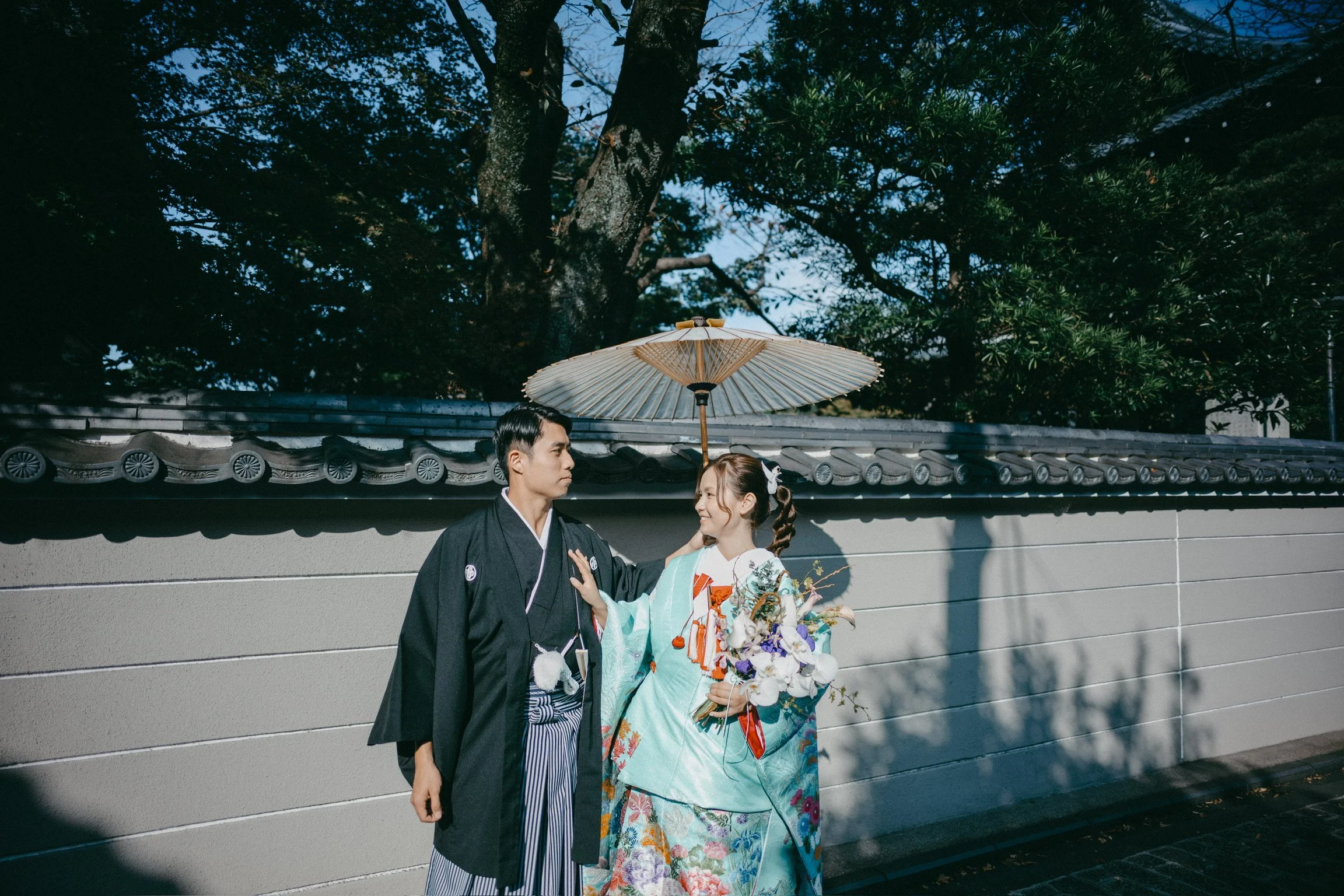 A young couple dressed in traditional Japanese wedding attire standing outside near a wall, the woman holding a bouquet of flowers and holding an parasol above their heads, with trees in the background.