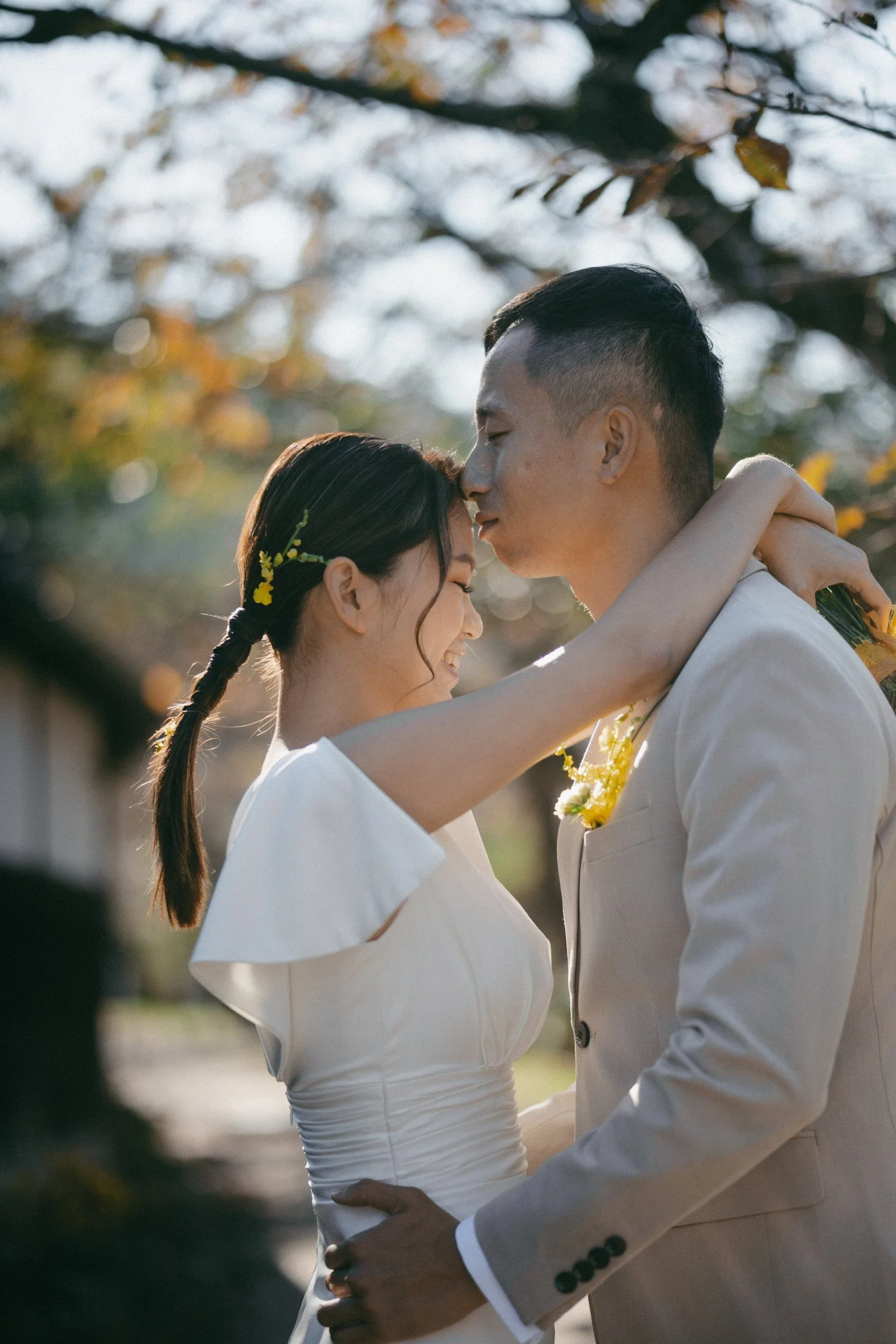 A couple in wedding attire sharing an intimate moment outdoors, with trees and sunlight in the background.