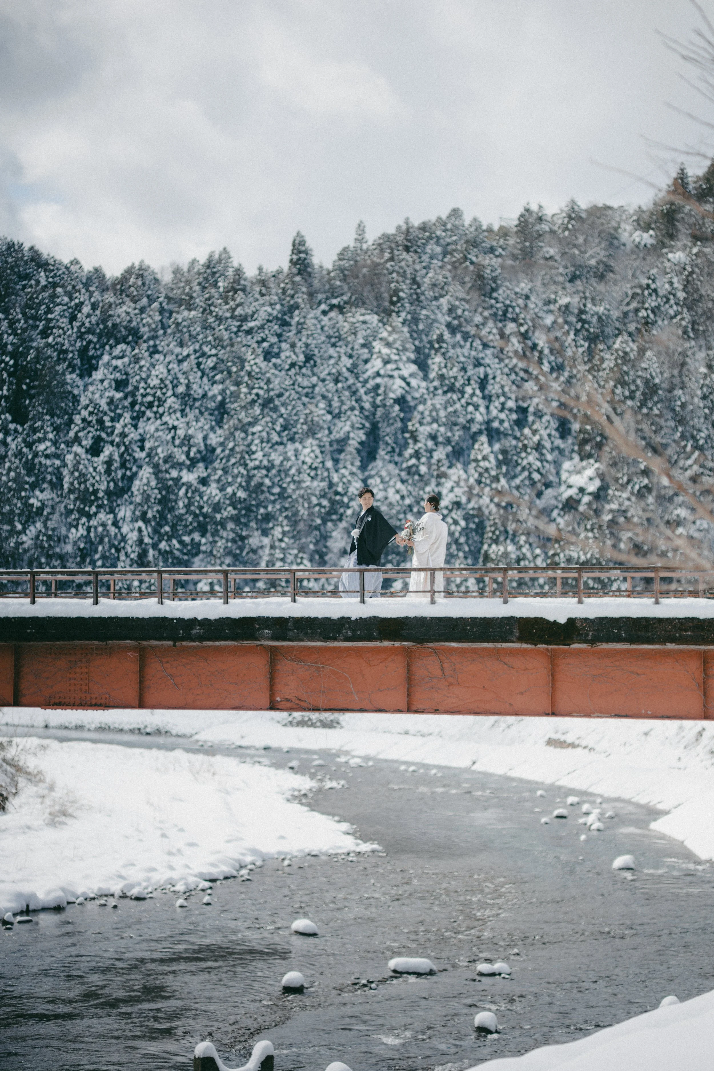 A couple dressed in traditional Japanese wedding attire standing on a snowy bridge over a river, with snow-covered trees in the background.