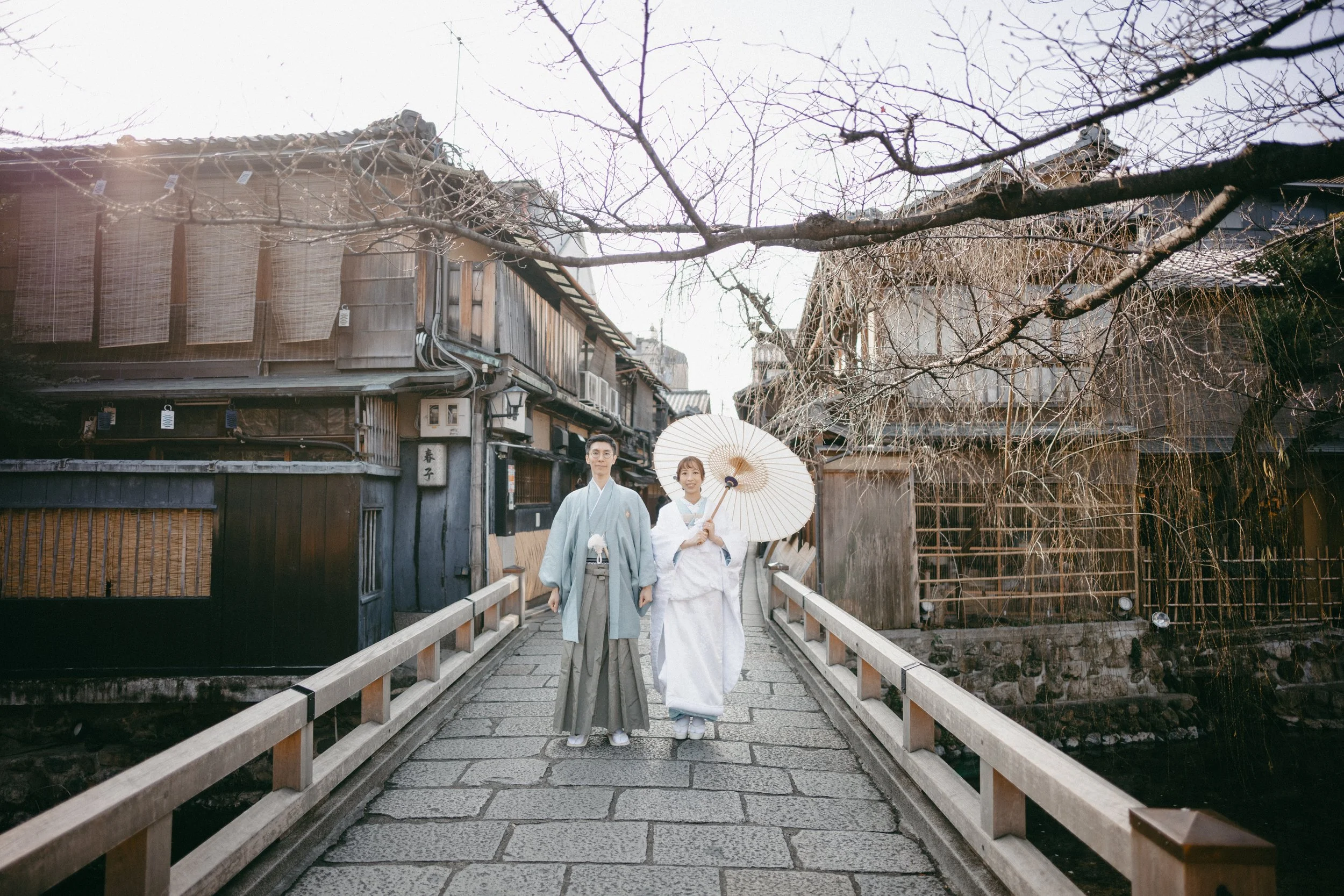 A couple dressed in traditional Japanese kimonos walking across a stone bridge in a historic town, with a woman holding a white parasol and a man with glasses.