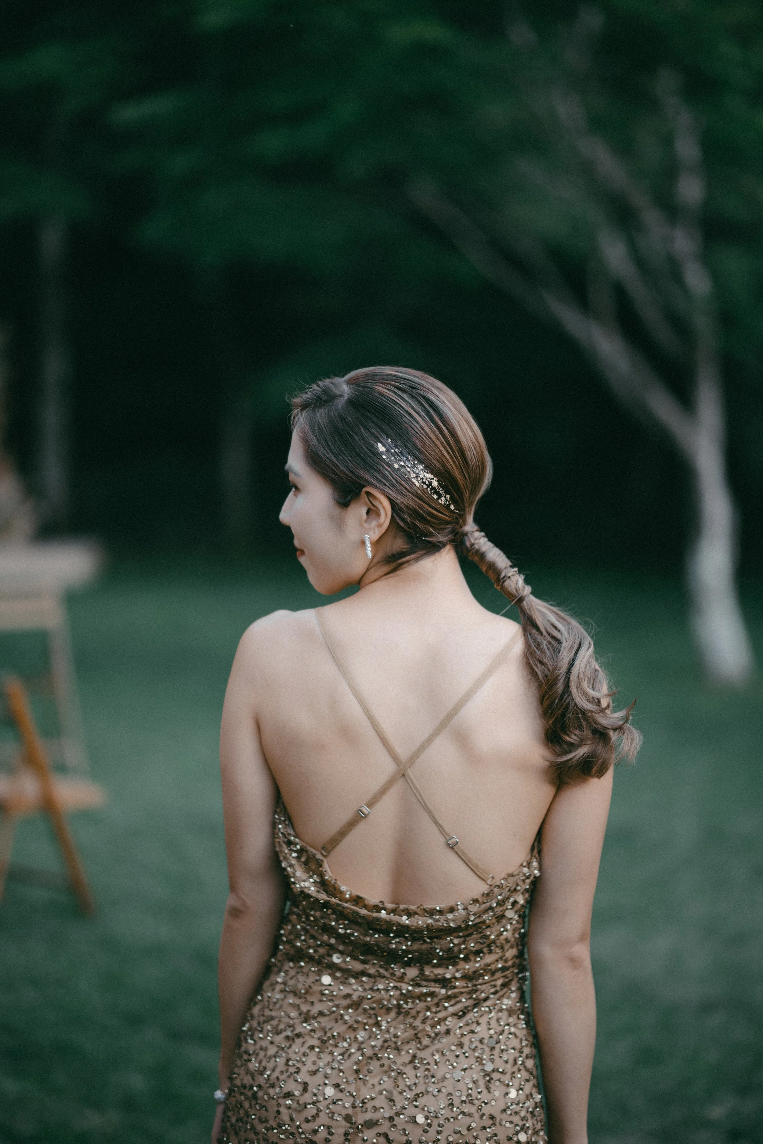 A woman with her back turned, wearing a glittery gold dress with thin straps and a rhinestone hair accessory, standing outdoors in a green park with trees.