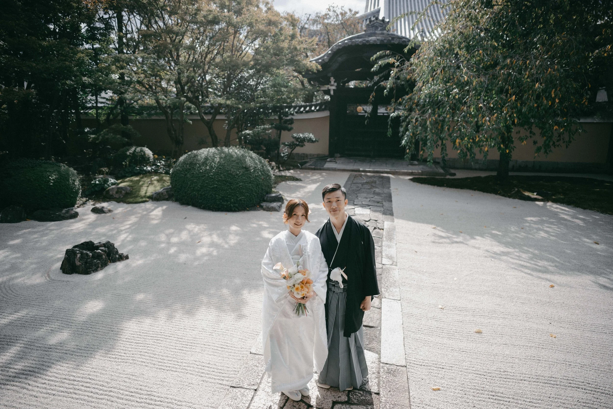 A bride and groom in traditional Japanese wedding attire standing on a monorail path in a Zen garden with rocks, trees, and a wooden gate in the background.