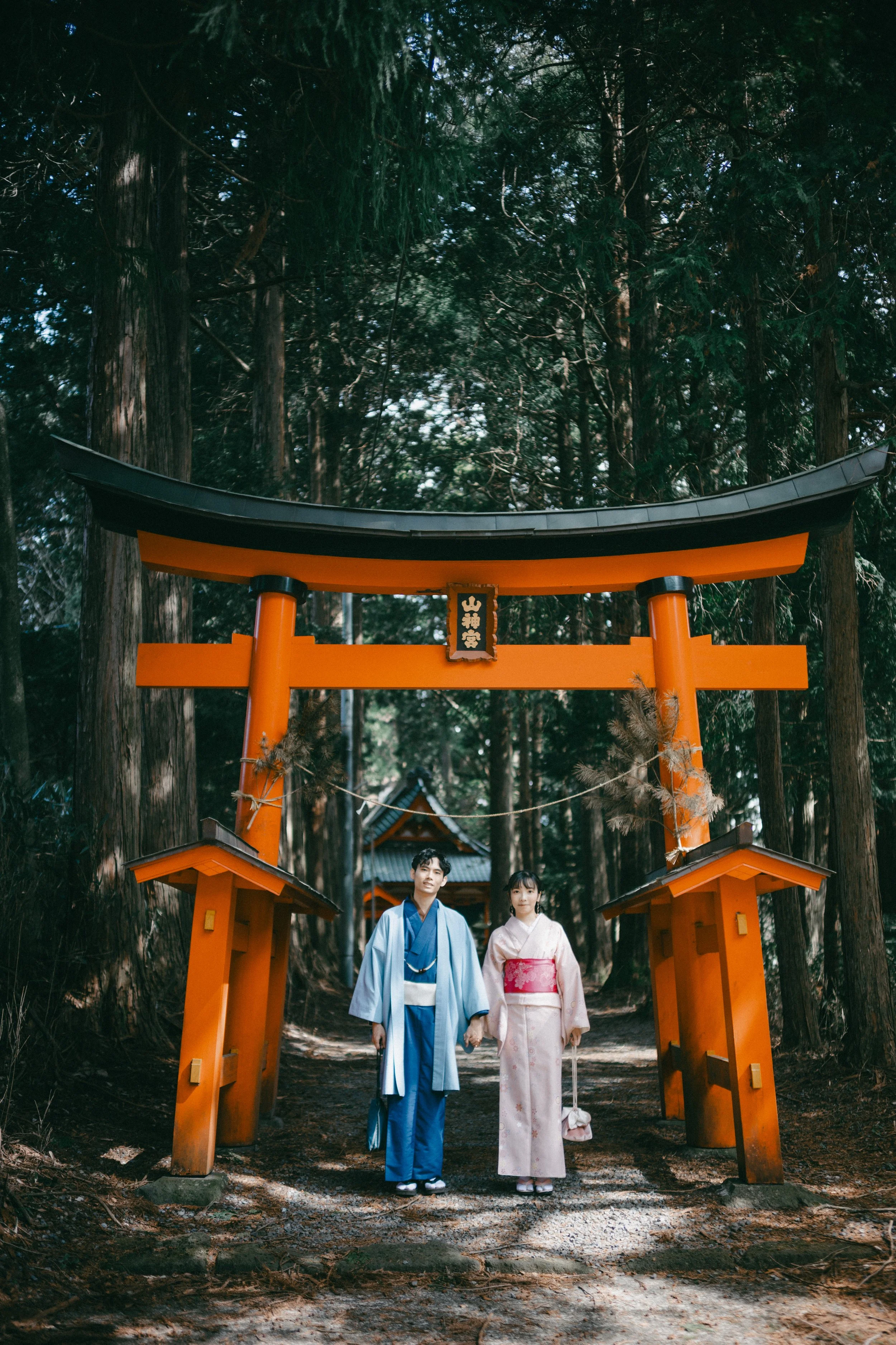 A man and woman in traditional Japanese attire standing under an orange torii gate in a wooded area.