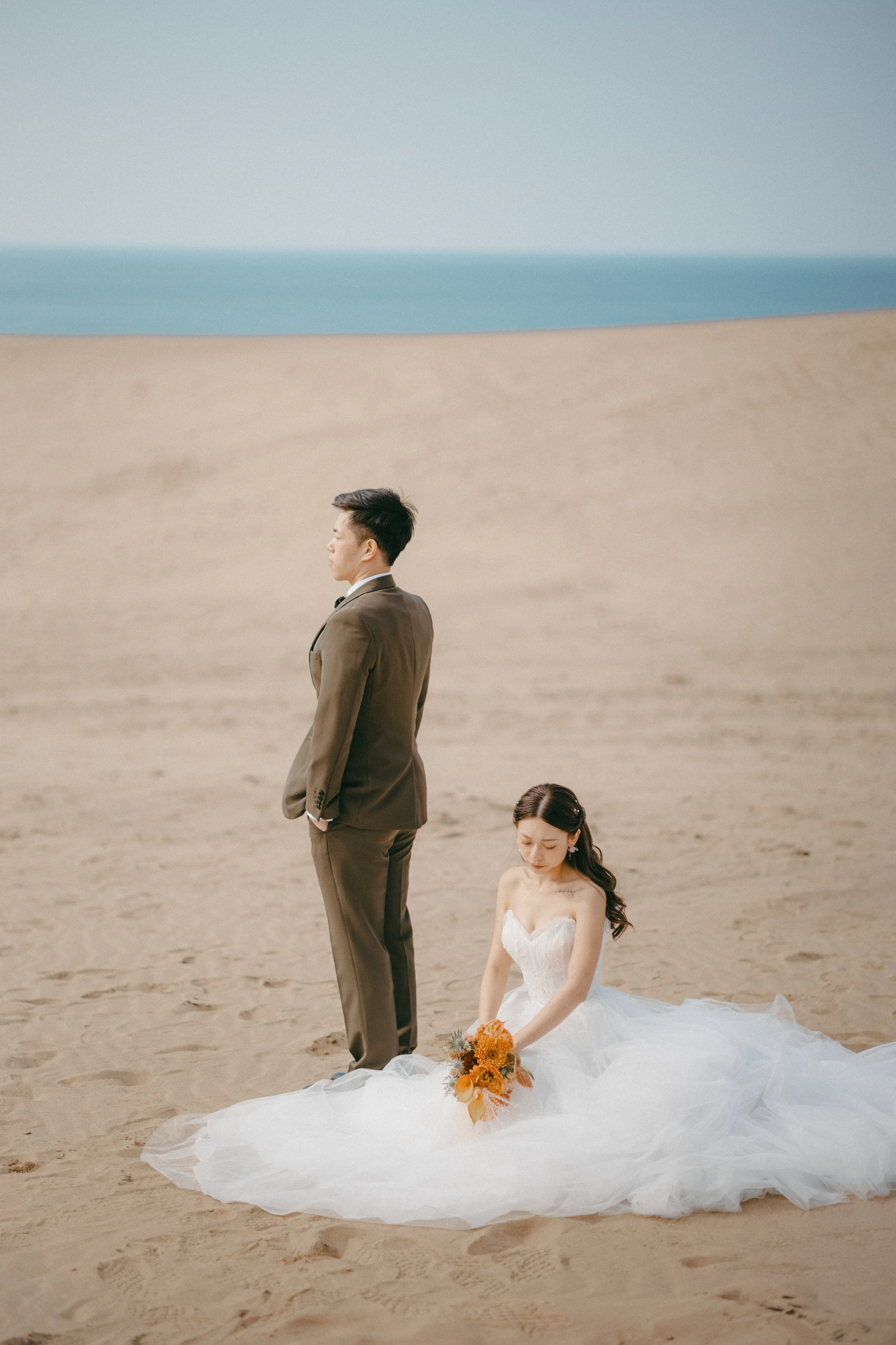 A bride in a white wedding dress sitting on the sand holding a bouquet, with a groom in a brown suit standing nearby on a beach.