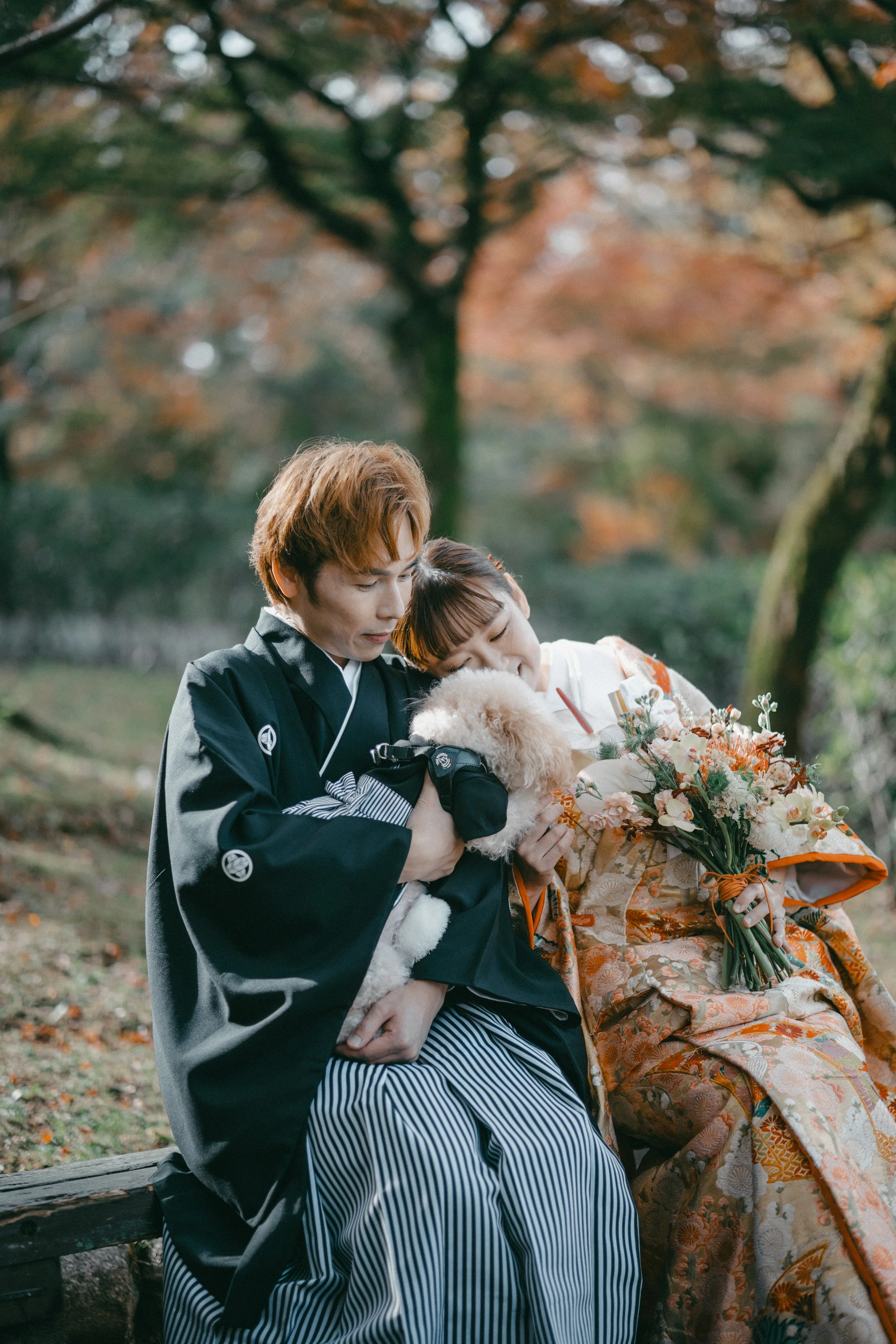 A couple dressed in traditional Japanese kimonos sitting outdoors on a park bench, holding a small white dog, with a woman holding a bouquet of flowers, surrounded by autumn trees.