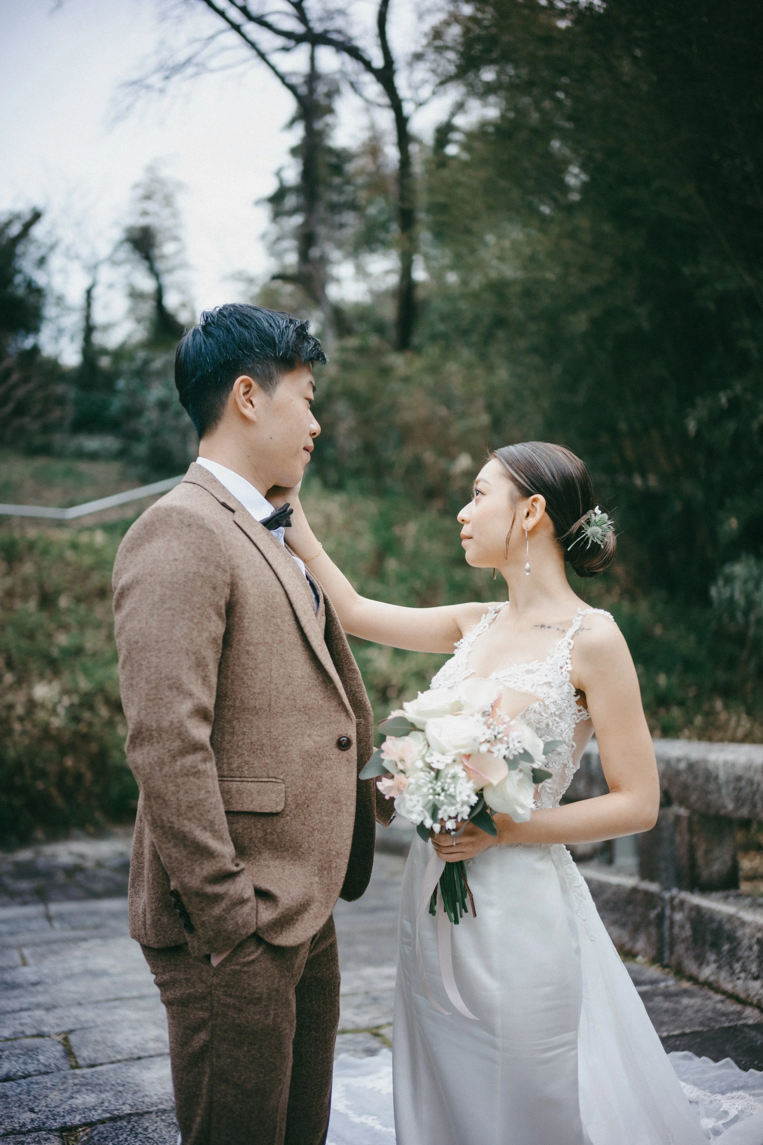A bride and groom stand close together outdoors during their wedding, with the bride holding a bouquet of white and blush roses and the groom wearing a brown suit, as the bride gently touches the groom's face.