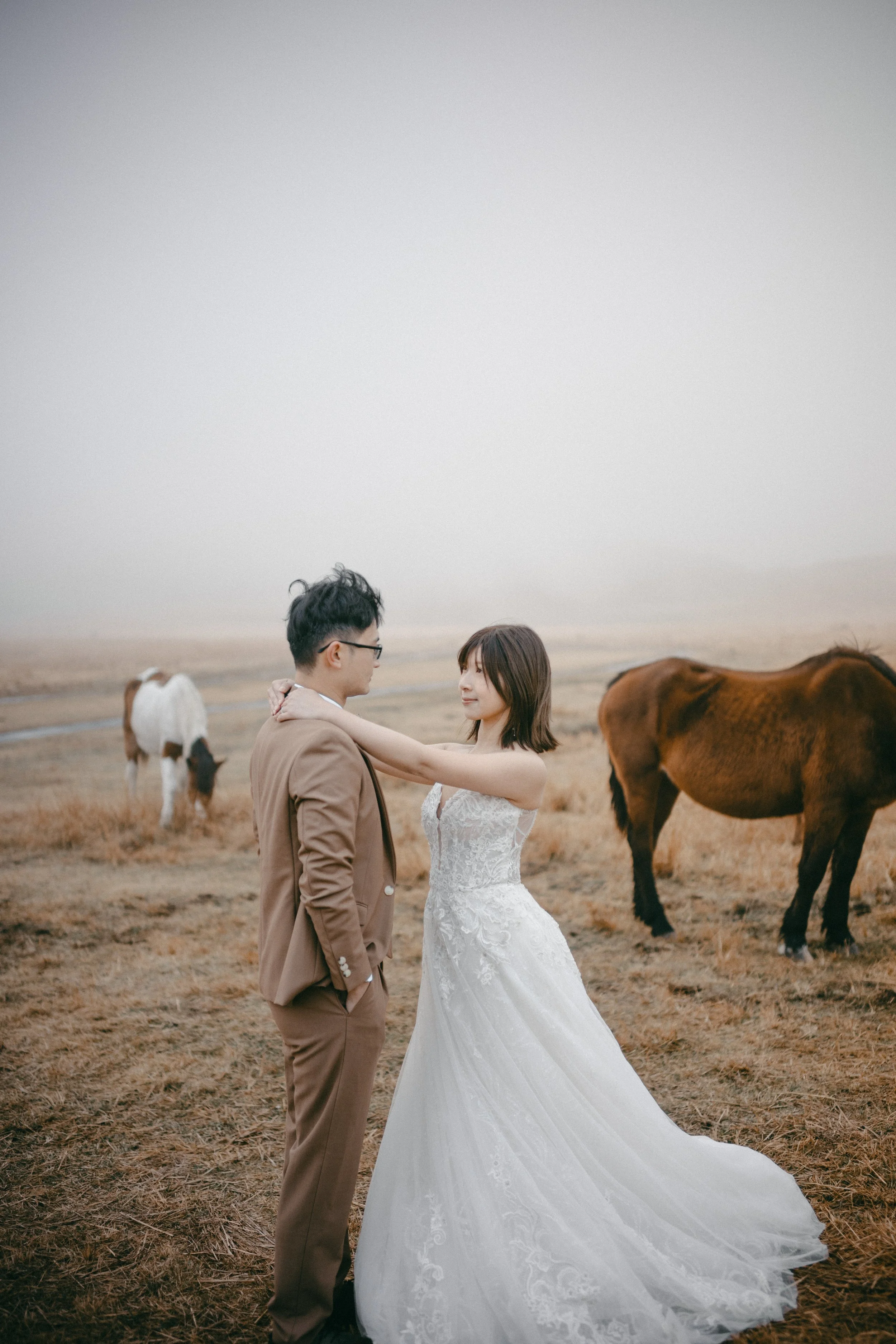 A bride and groom standing close together in a field with horses, the bride in a white wedding dress and the groom in a brown suit, outdoors on a foggy day.