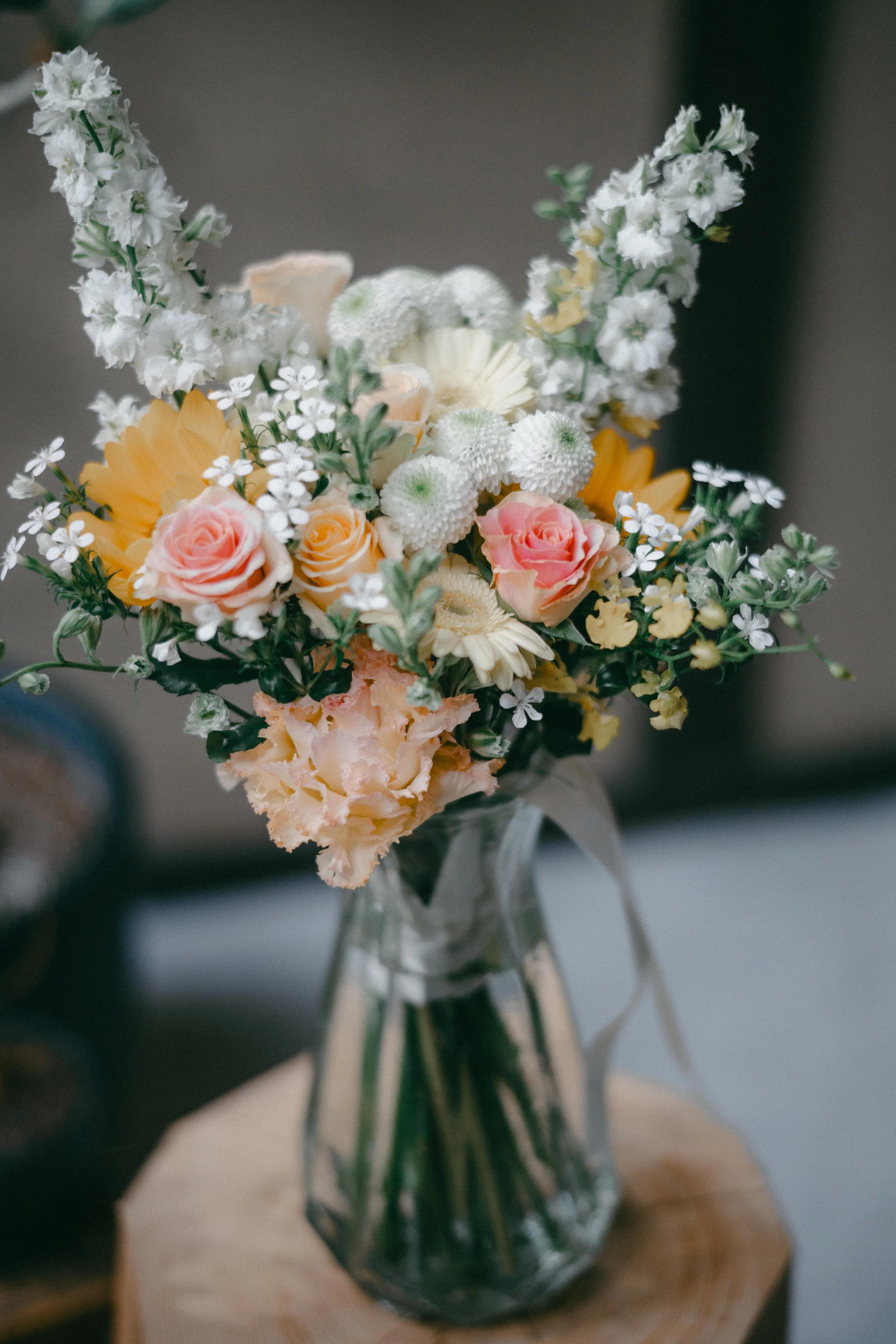 A bouquet of mixed flowers including pink roses, white daisies, white chrysanthemums, and white snapdragons in a clear glass vase on a wooden surface.