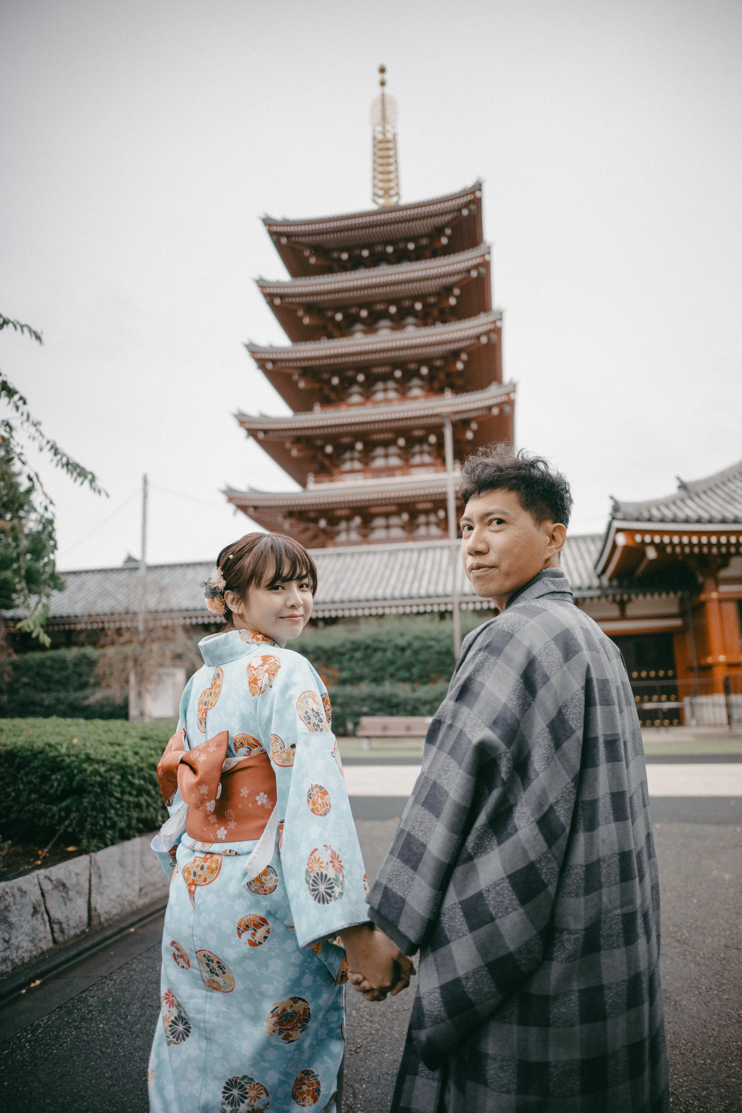 A couple holding hands and looking back at the camera, standing in front of a traditional Japanese five-story pagoda, wearing yukatas, in an outdoor setting.
