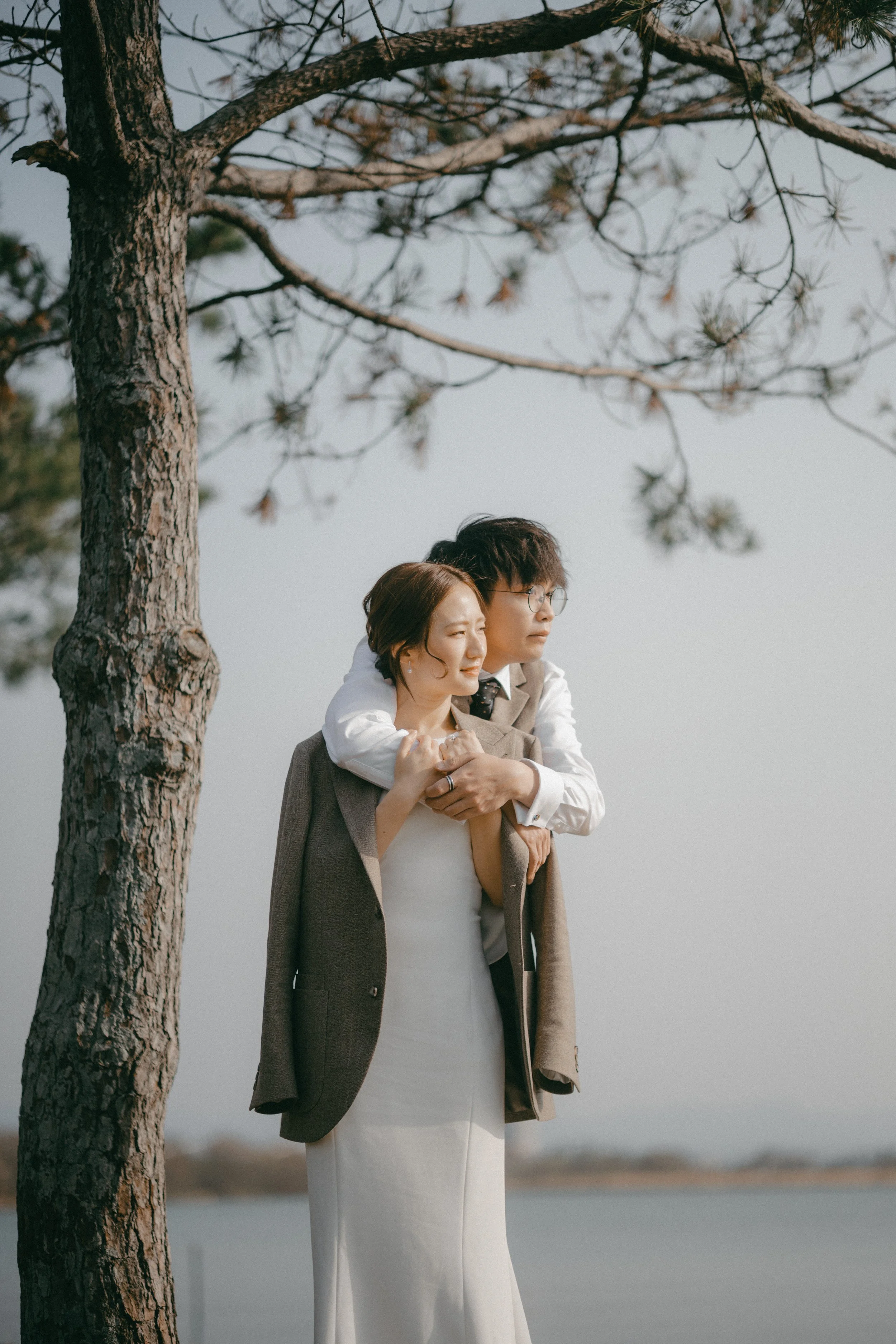 A couple dressed in formal attire outdoors by a tree, with the man giving the woman a piggyback ride, both smiling and enjoying a scenic view of water in the background.