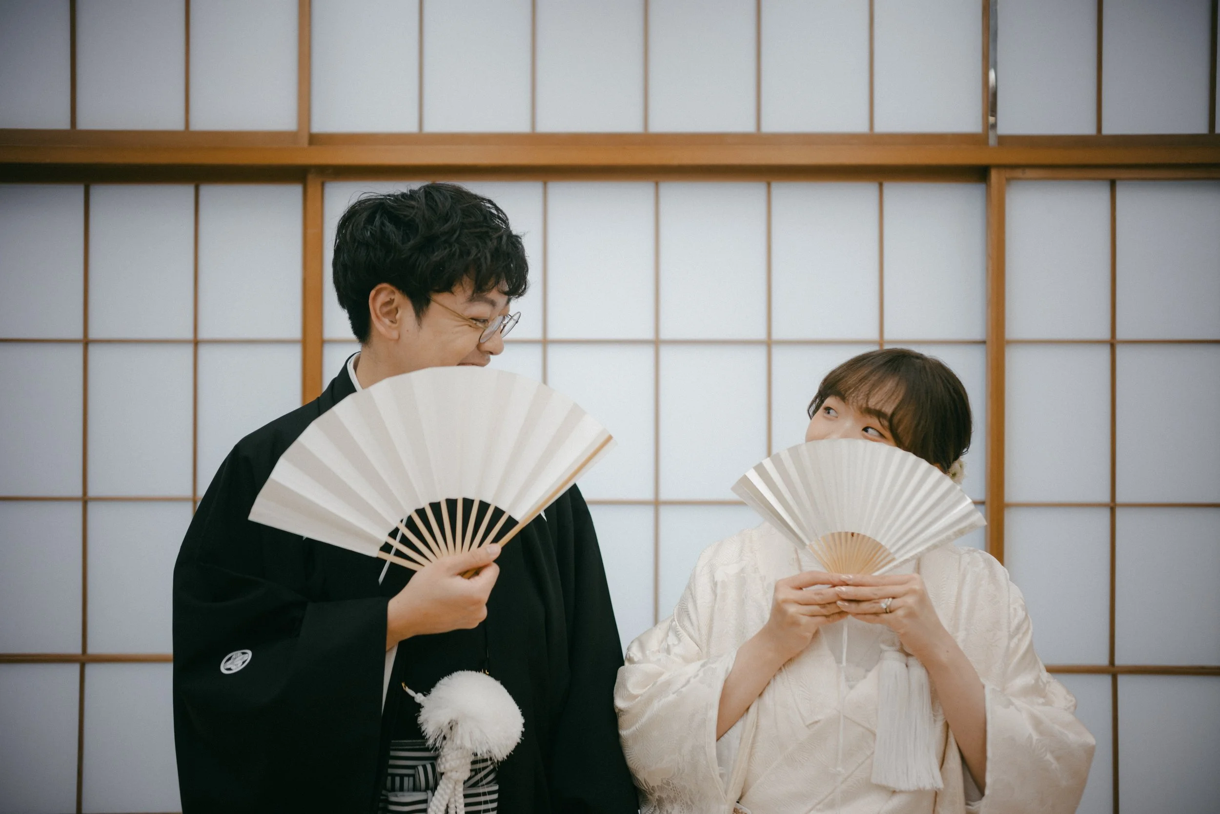 A man and a woman wearing traditional Japanese wedding attire holding fans and smiling at each other in front of a shoji screen.