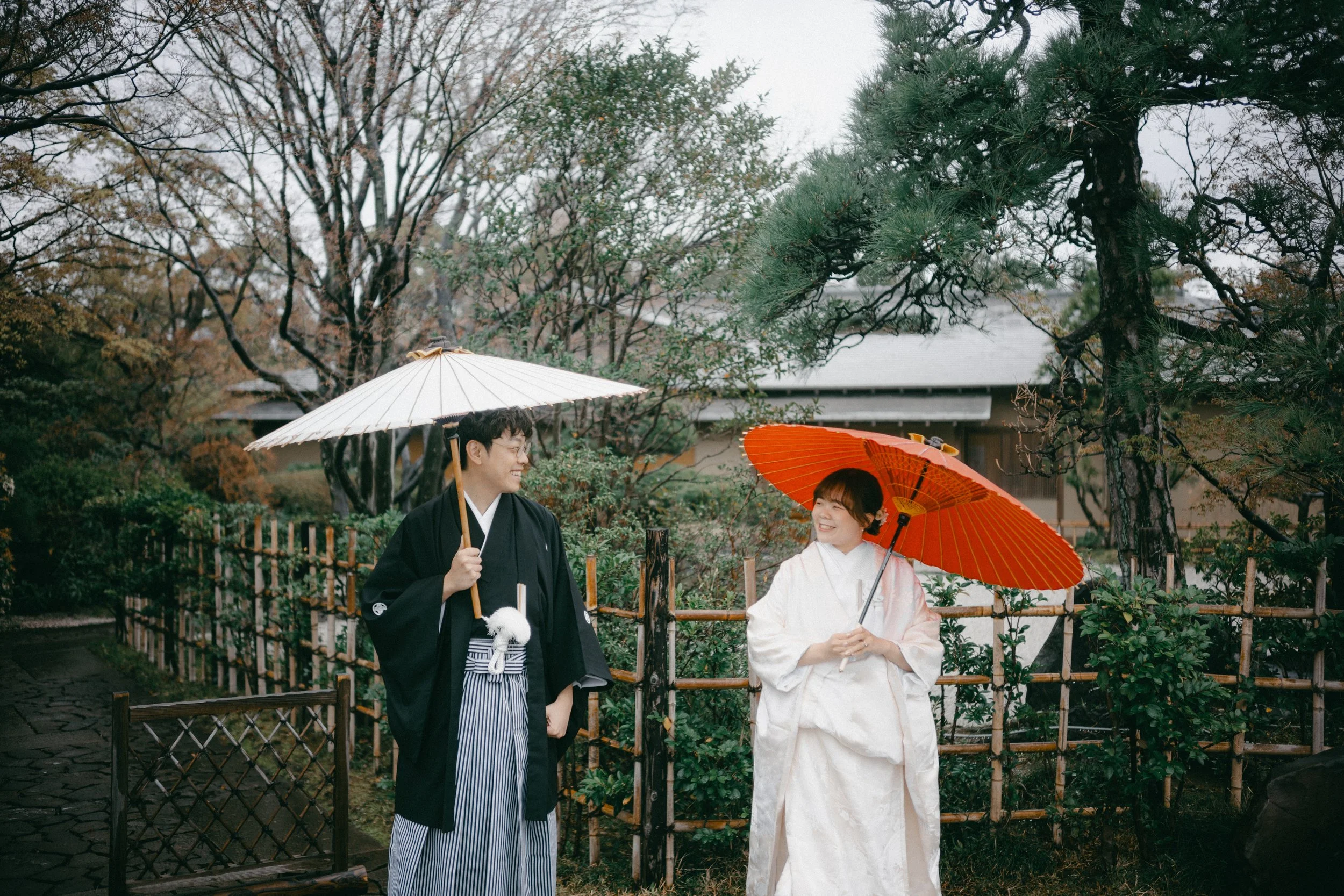 A couple dressed in traditional Japanese wedding attire holding umbrellas and smiling at each other in a garden.