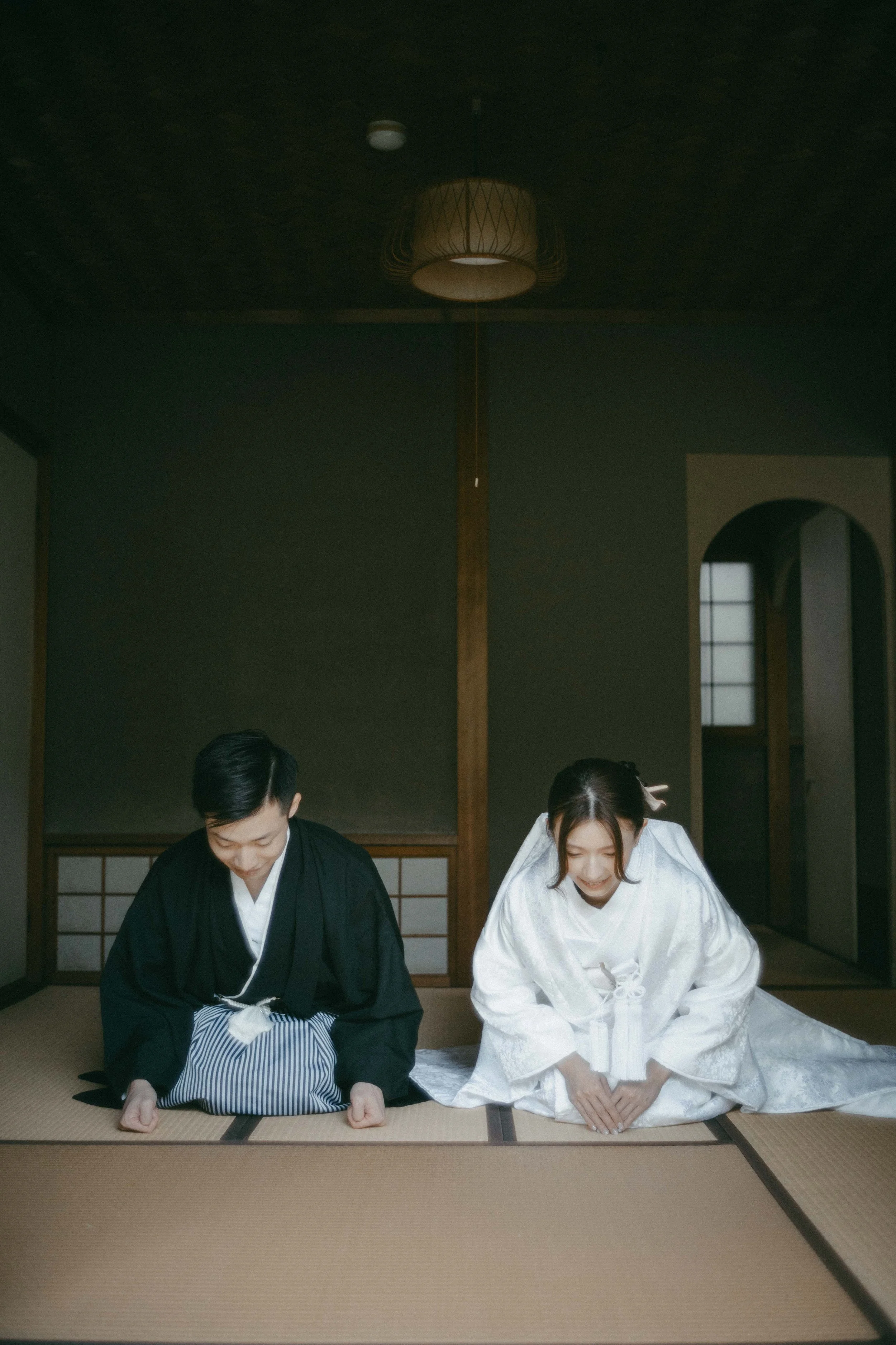 A couple dressed in traditional Japanese wedding attire bowing on tatami mats inside a Japanese-style room.