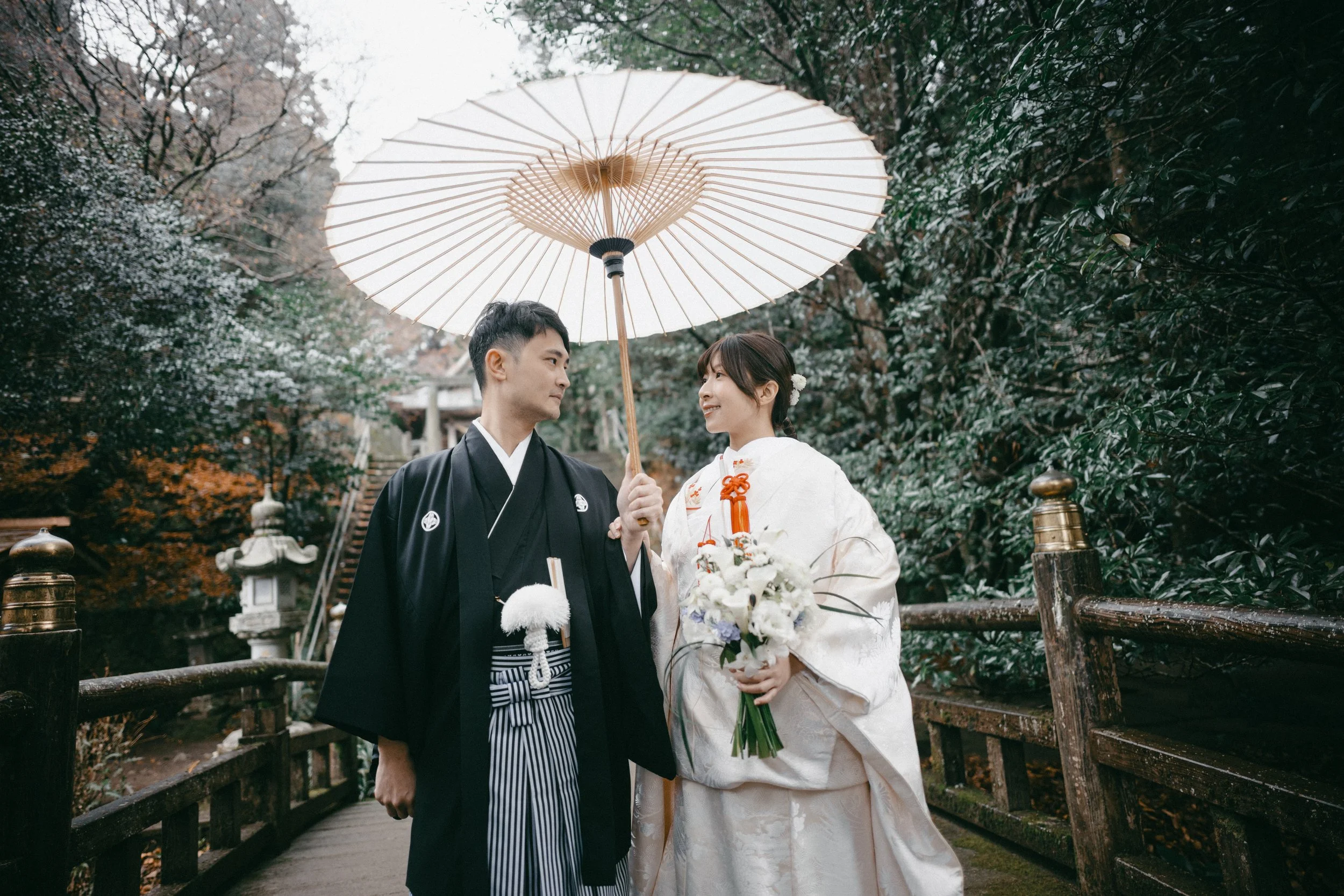 A Japanese bride and groom in traditional wedding attire standing on a wooden bridge outdoors, sharing a moment under a large paper umbrella, surrounded by lush trees.