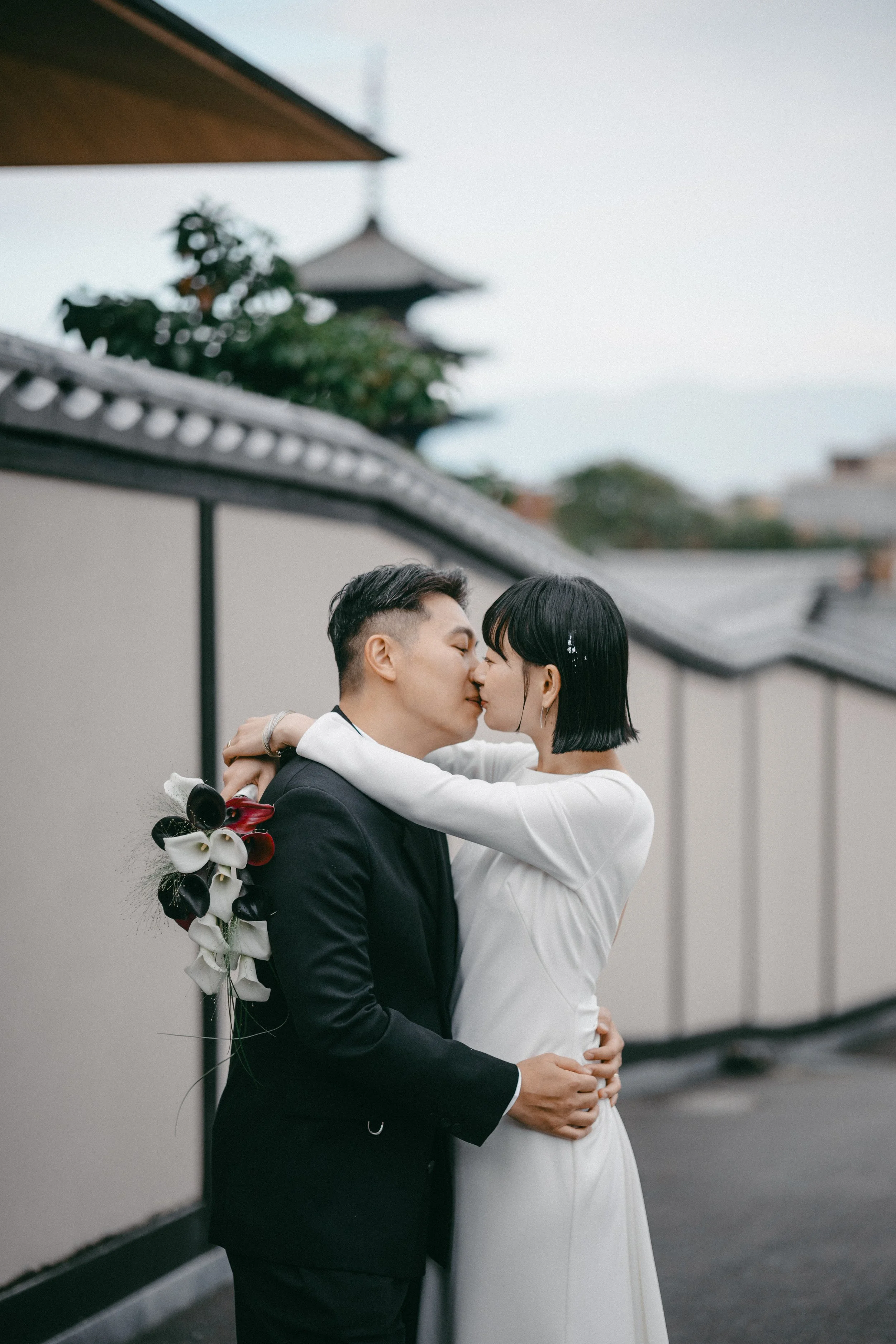 A couple kissing, with the woman wearing a white dress and the man in a black suit, holding a bouquet of flowers, outdoors in front of traditional Asian-style rooftops.