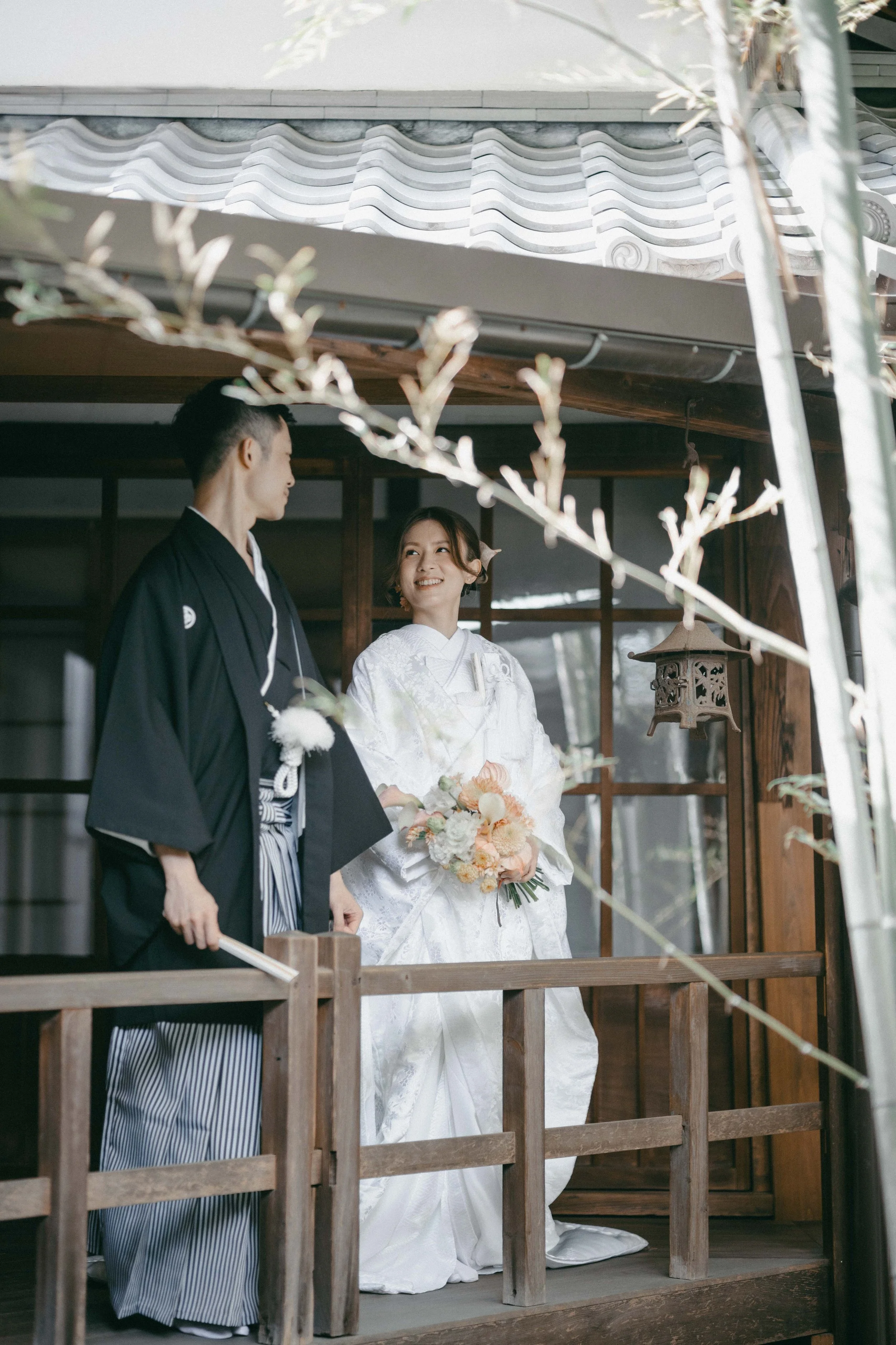 A couple dressed in traditional Japanese wedding attire, standing on a wooden porch and smiling at each other. The woman is wearing a white wedding kimono holding a bouquet, and the man is wearing a black kimono with striped hakama.