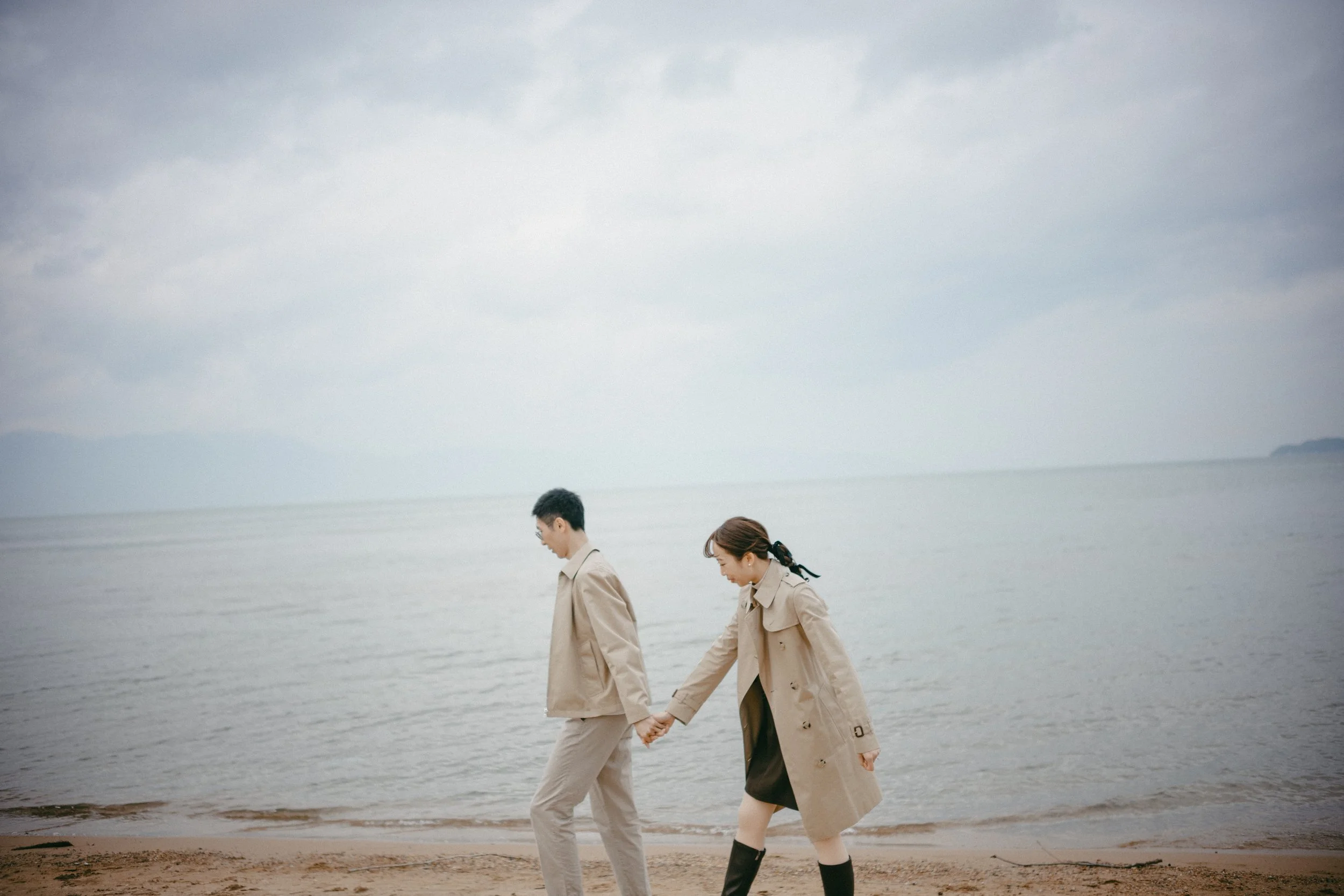 A couple holding hands walking along a beach with an overcast sky.