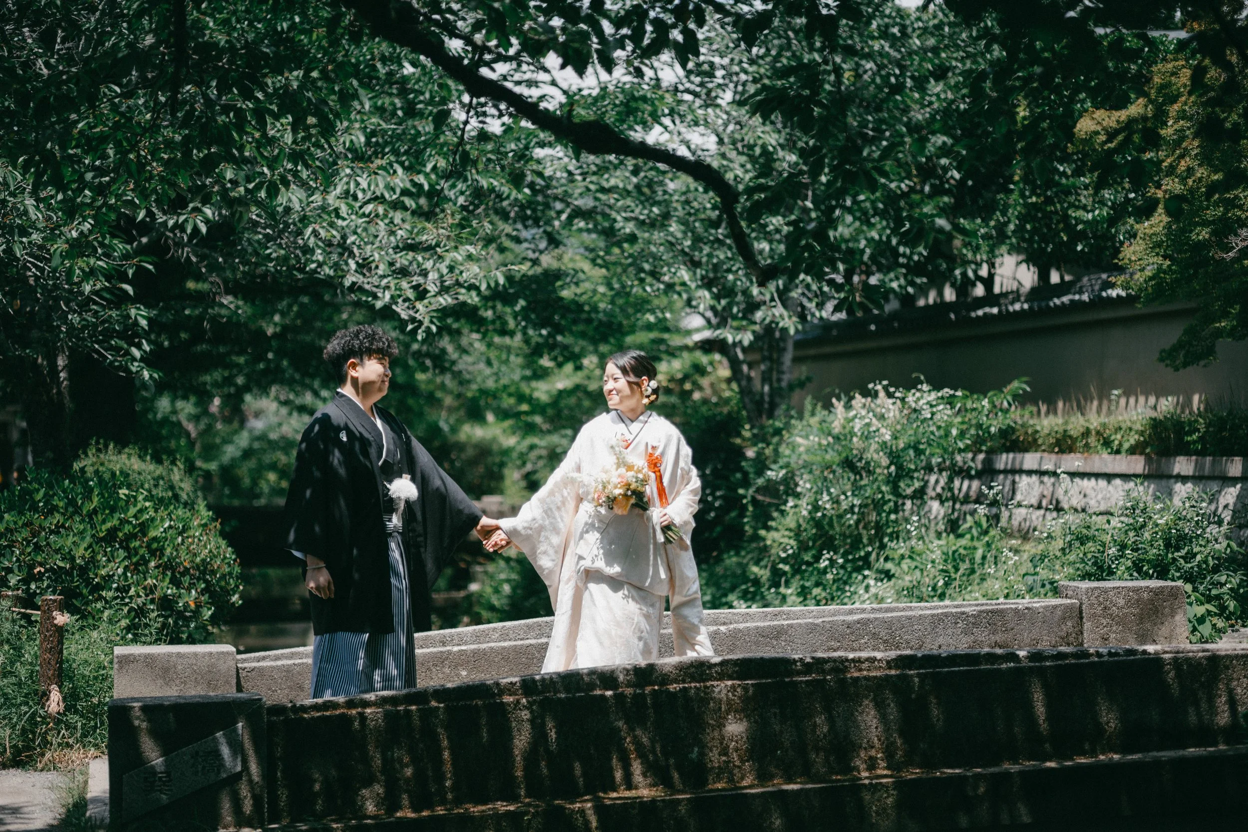 A couple in traditional Japanese wedding attire holding hands and smiling at each other in a lush green outdoor setting.