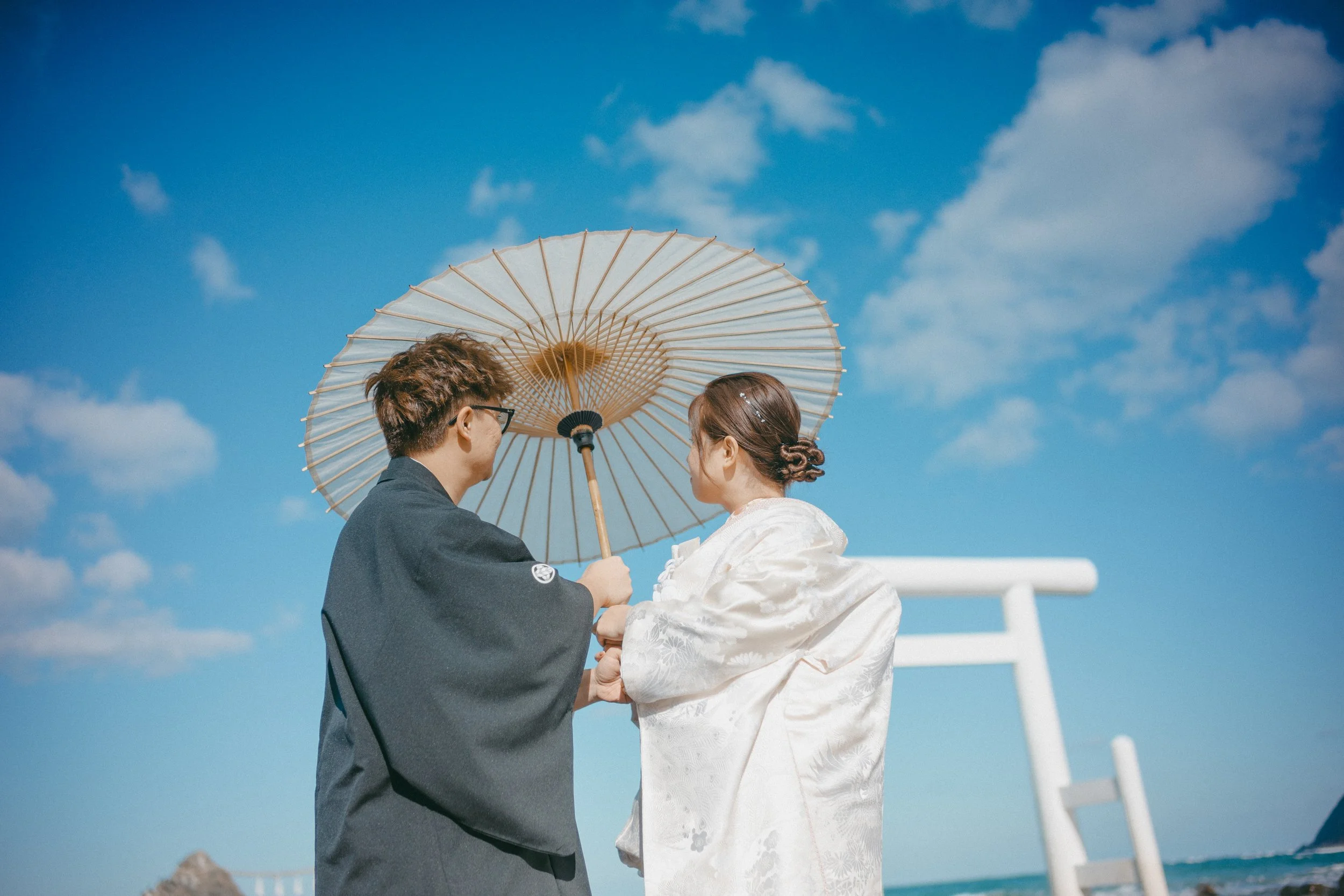 A couple dressed in traditional Japanese wedding attire holding hands under a large parasol on a sunny beach with a blue sky and some clouds.